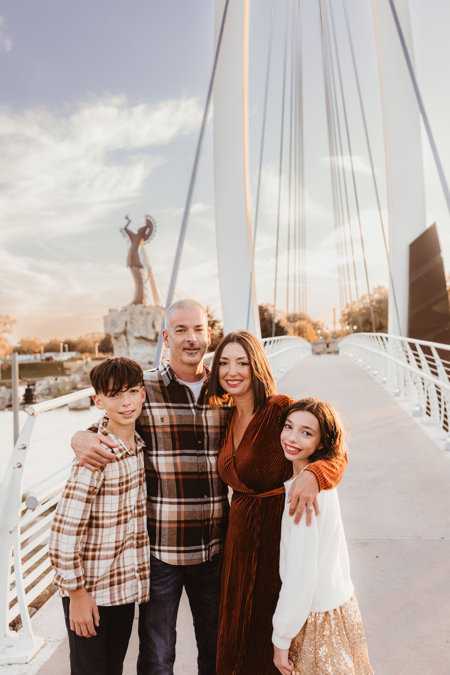 Family of four poses on modern suspension bridge at sunset in urban setting.
