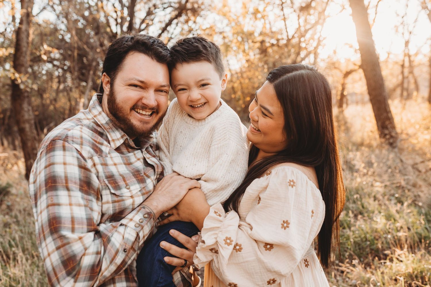 Happy family of three laughs together outdoors in golden autumn field at sunset.