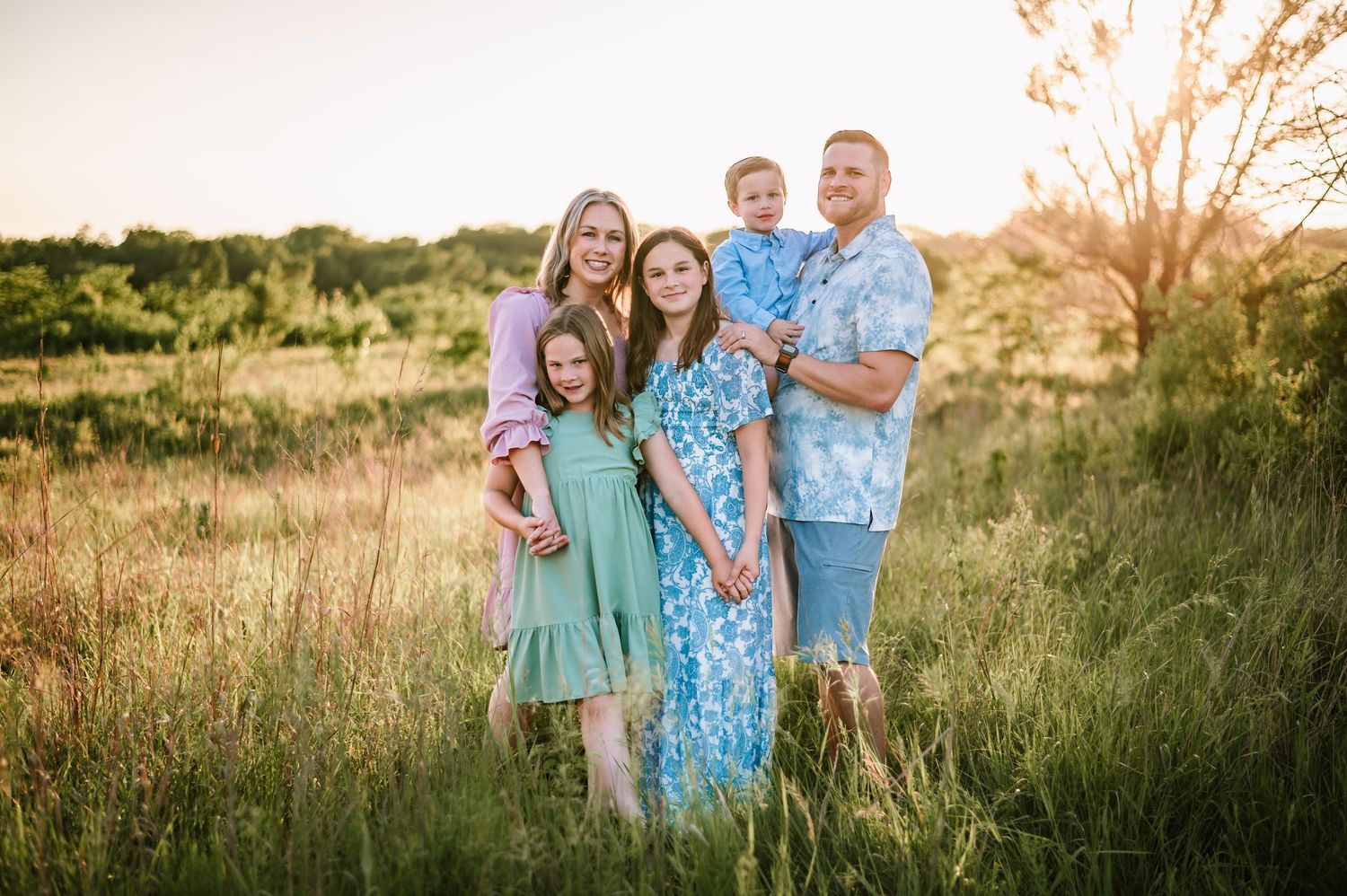 Happy family of five posing together in a sunlit green meadow at golden hour.