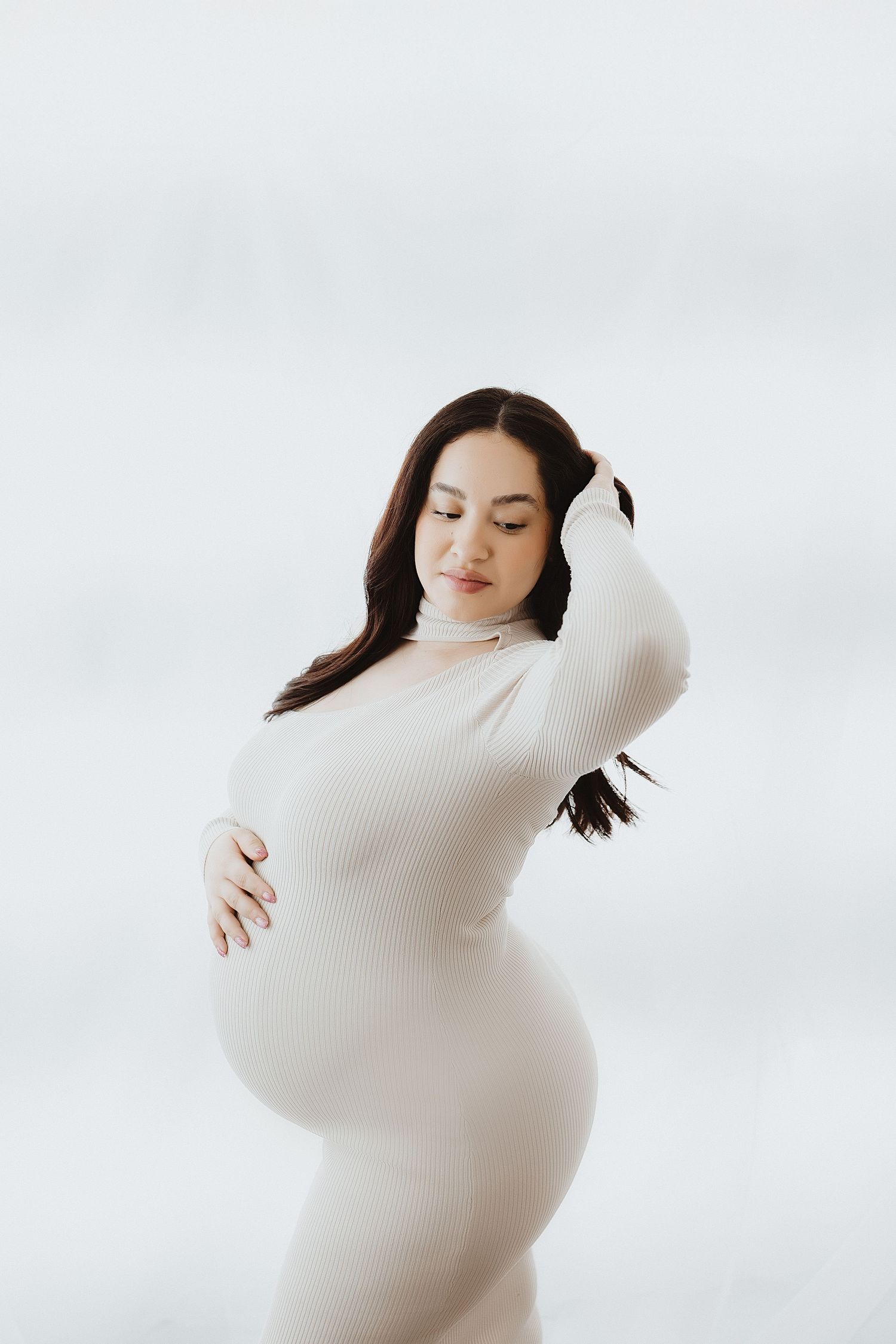 Elegant pregnant woman in fitted white ribbed dress cradles her baby bump in a serene studio portrait.