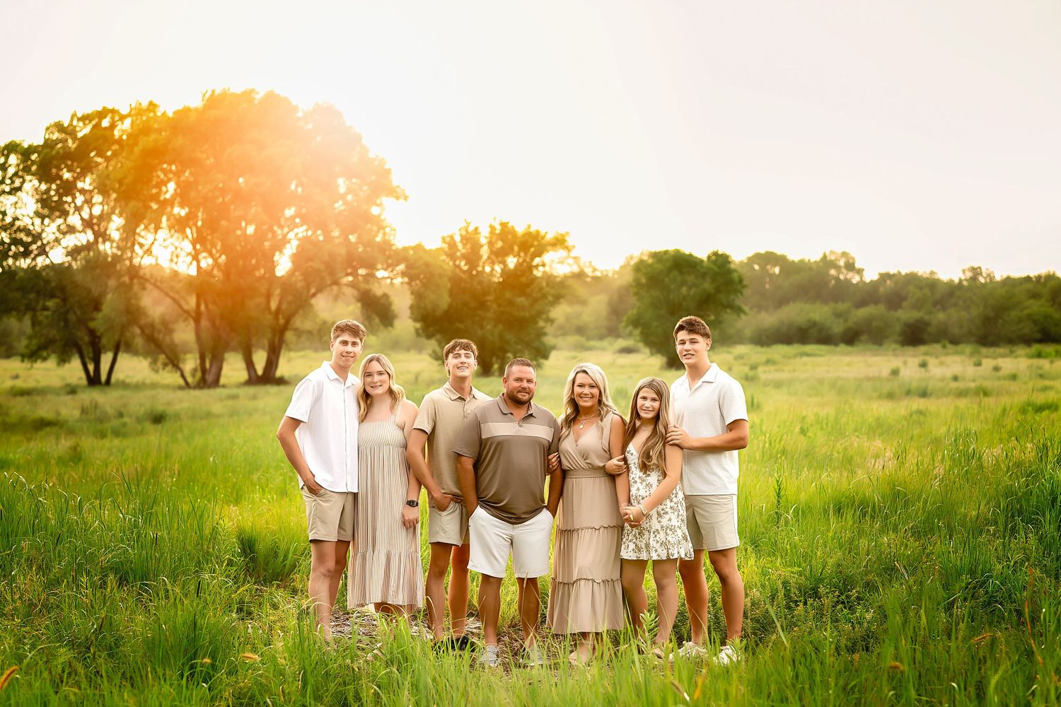 Family of seven posing together in a sunlit green meadow during golden hour, wearing coordinated neutral and white outfits.