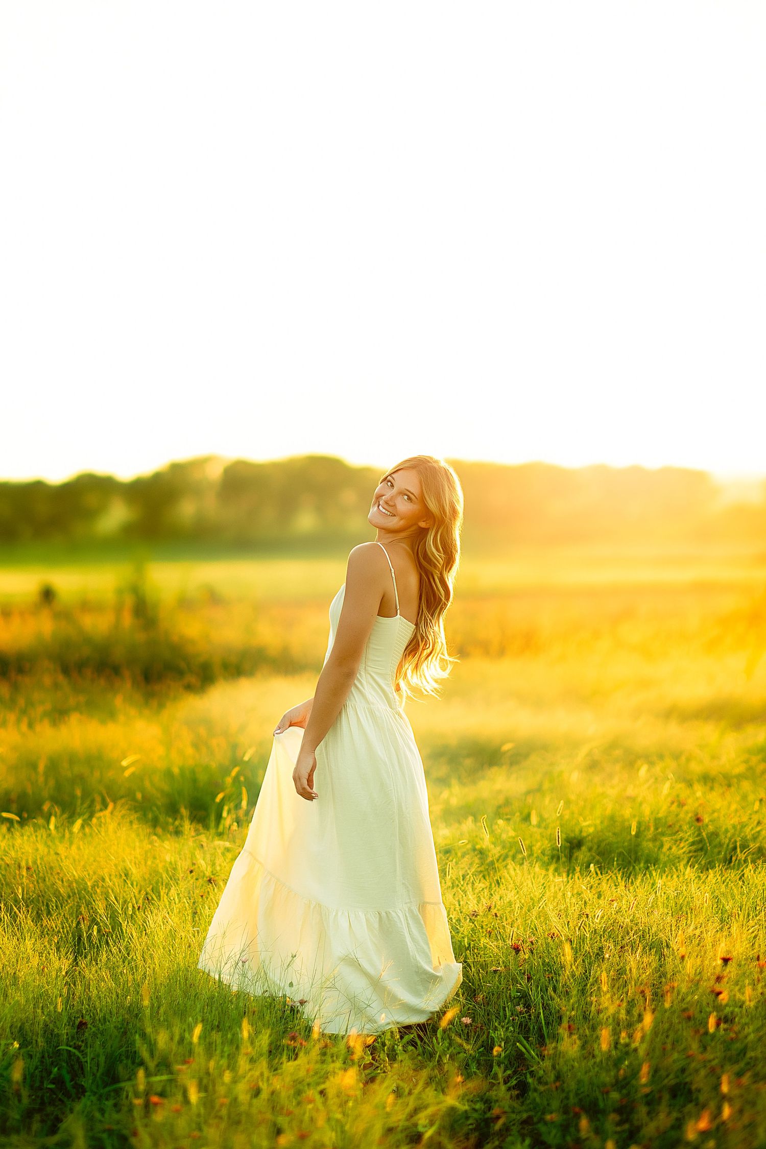 Young woman in white dress standing in sunny green field at golden hour, smiling over shoulder.