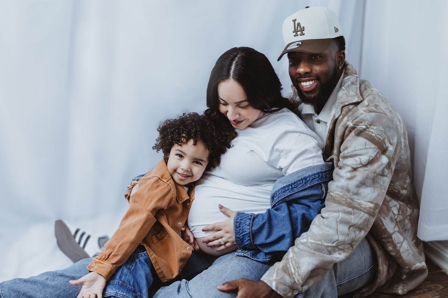 A smiling military father and pregnant mother cuddle their happy toddler in a warm family photo.
