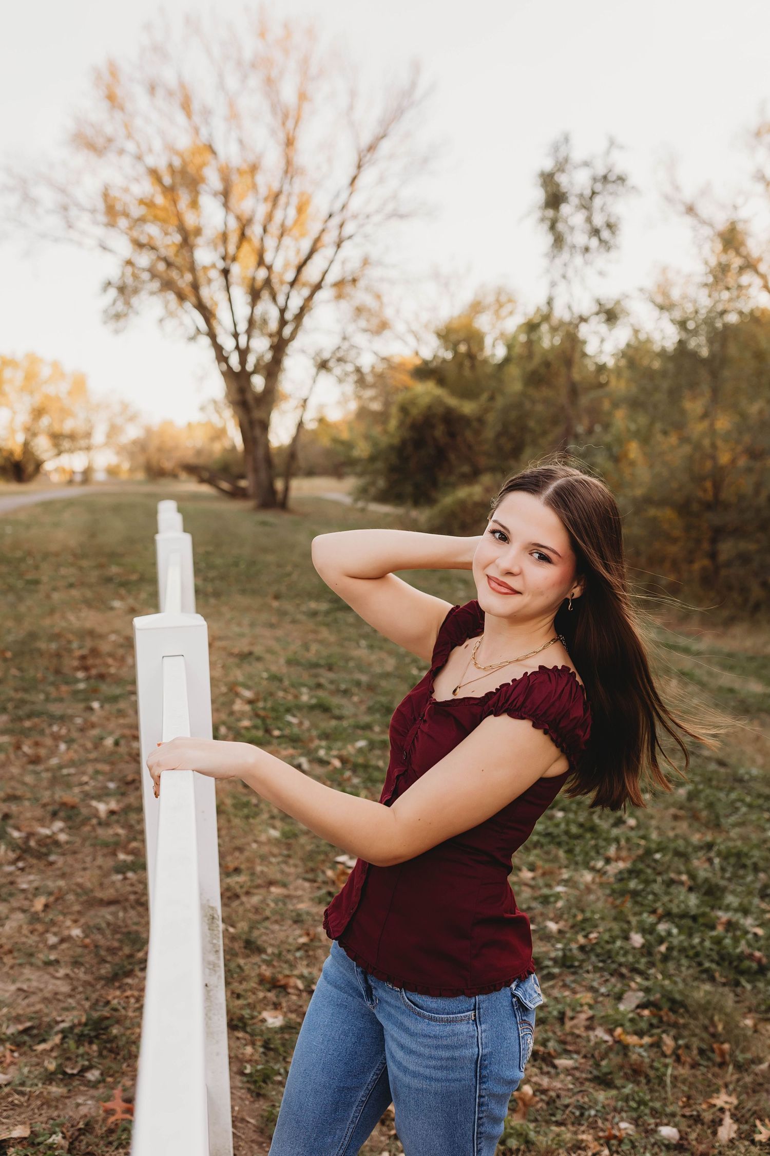 Young woman in burgundy top and jeans poses by white fence post in autumn park setting.