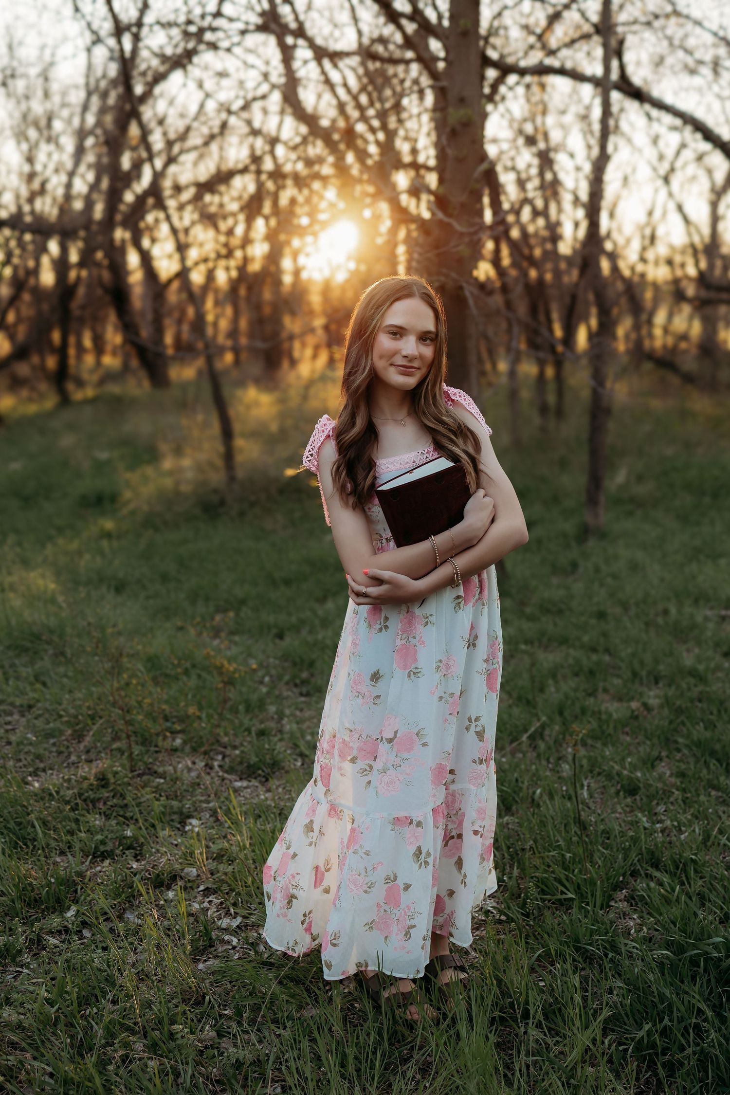 Young woman in floral maxi dress holding a book outdoors at golden sunset in a wooded garden setting.