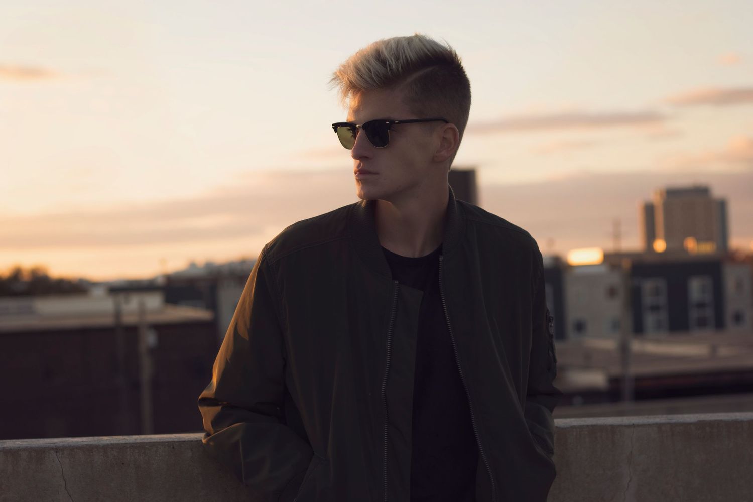 Stylish young man in sunglasses and bomber jacket posing on rooftop at sunset.
