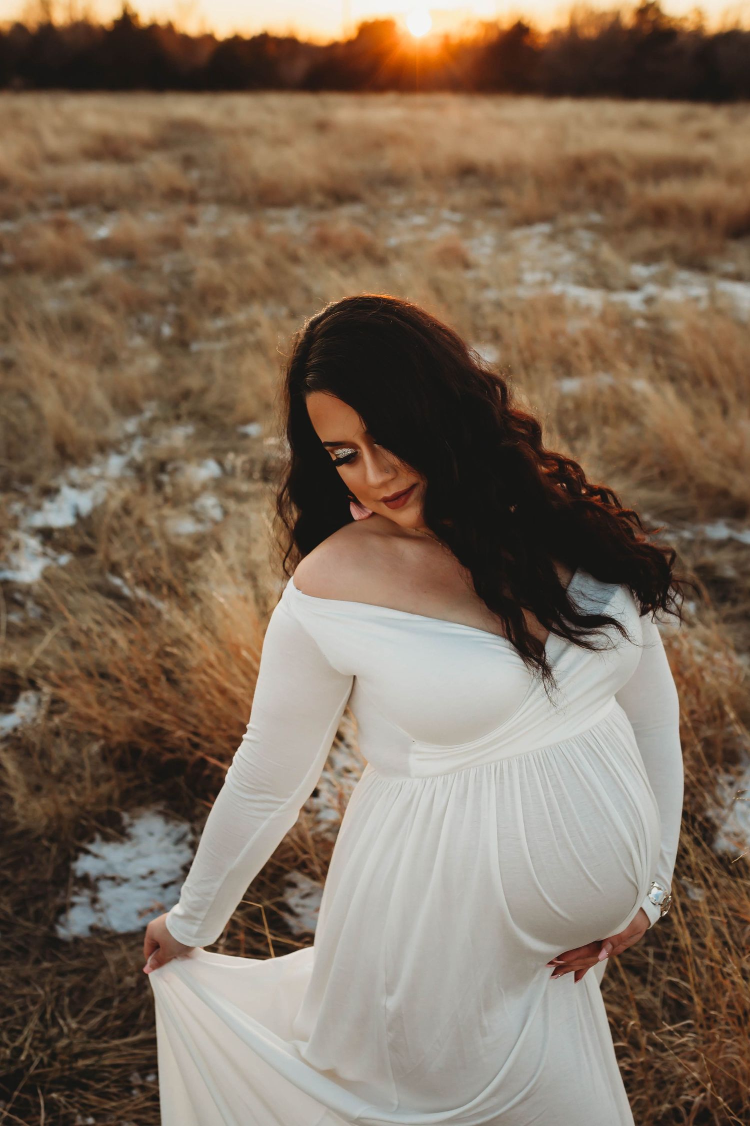 A pregnant woman in a flowing white off-shoulder gown poses gracefully in a golden field at sunset.