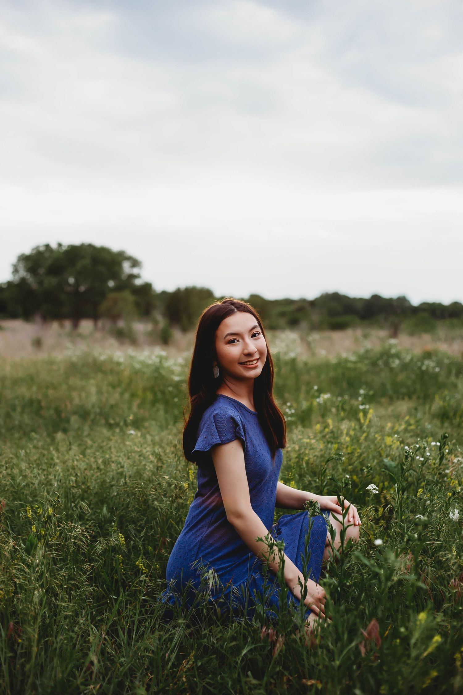 Young woman in blue dress sitting in a wildflower field, smiling over her shoulder on a cloudy day.