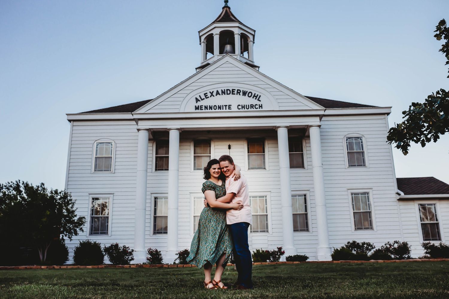Couple embracing in front of Alexanderwohl Mennonite Church at dusk.