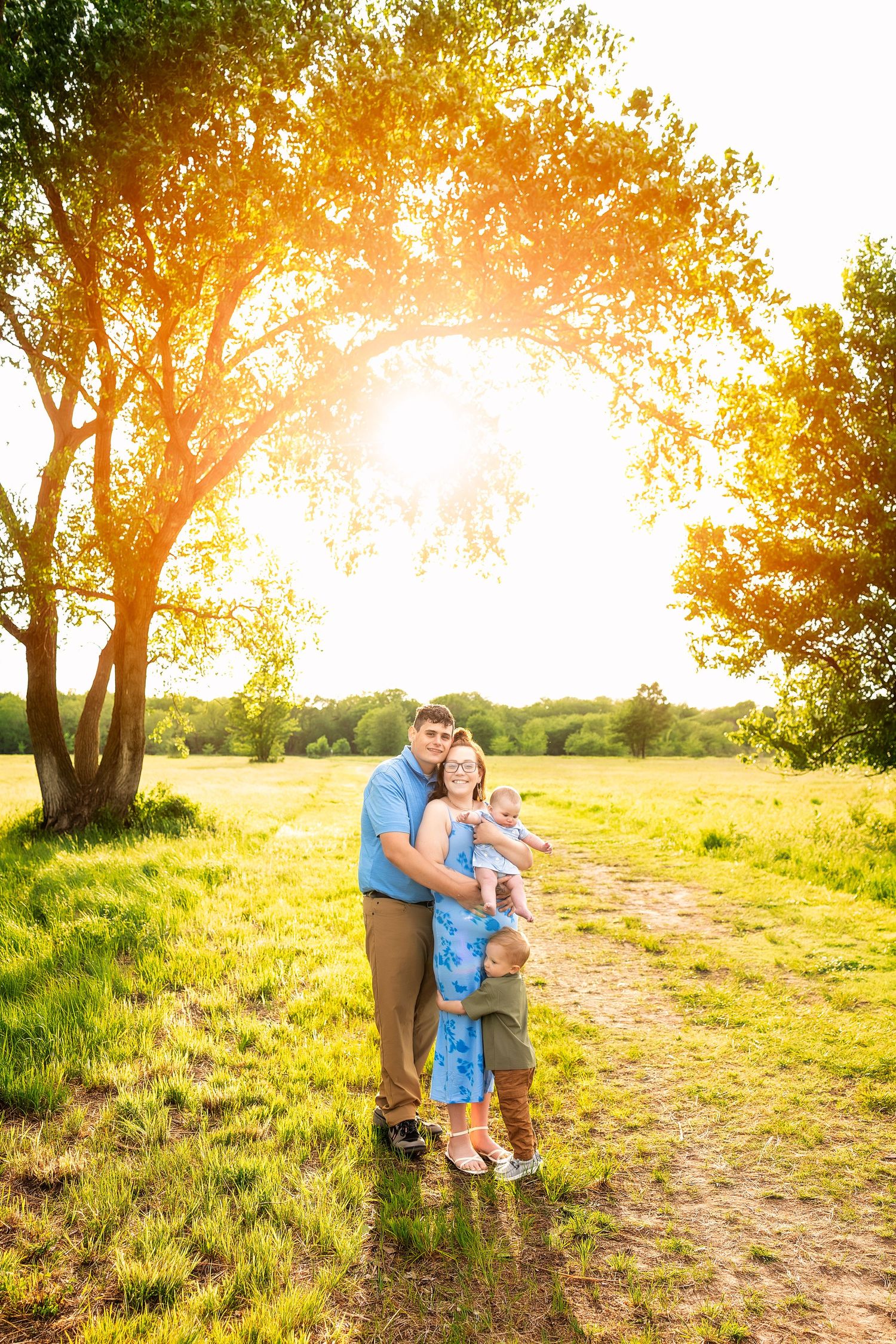 Family of three posing on a sunny dirt path surrounded by lush green trees and golden sunlight.