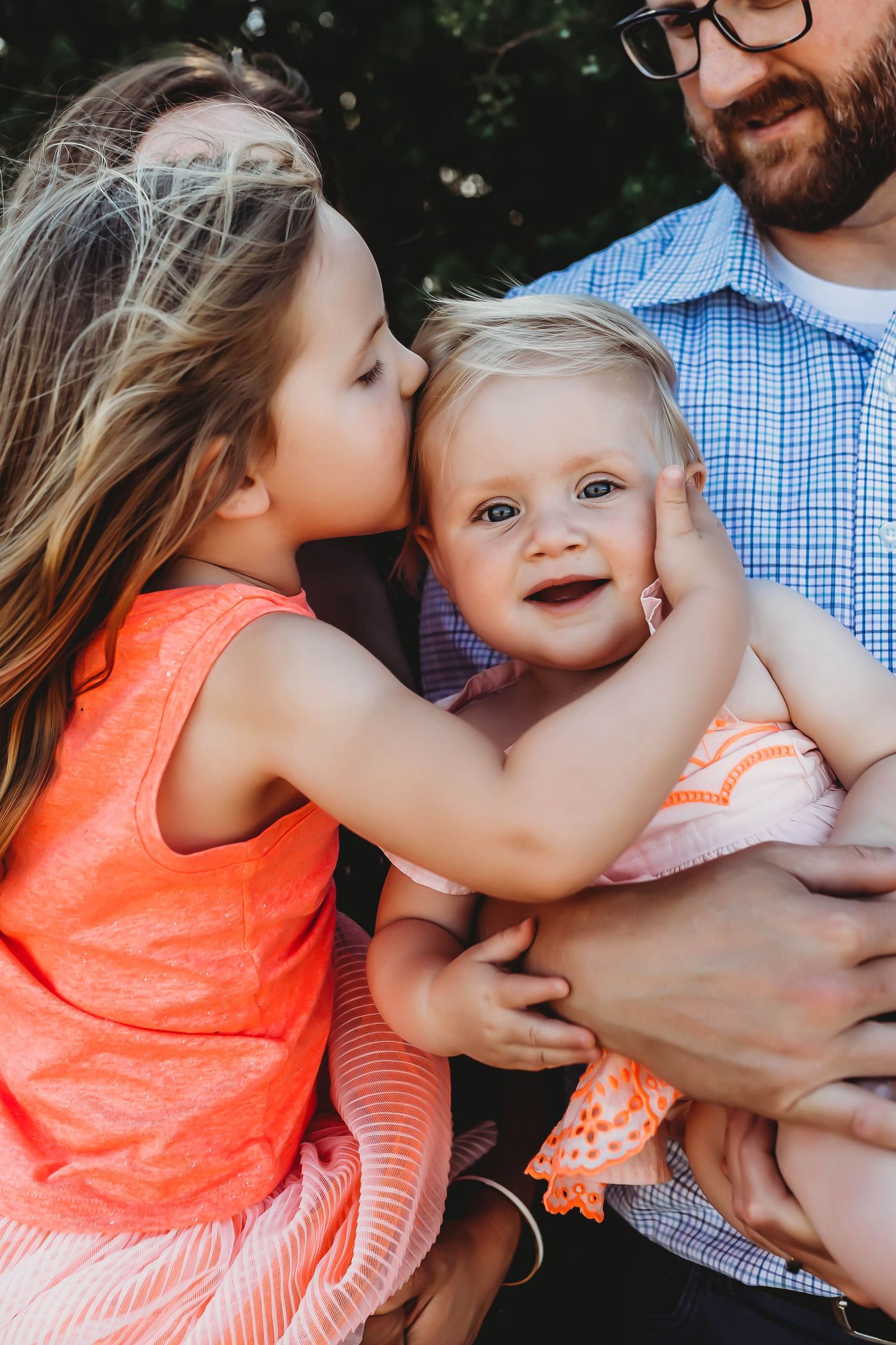 Sweet older sister kissing baby sibling on the cheek while loving father holds the baby outdoors.