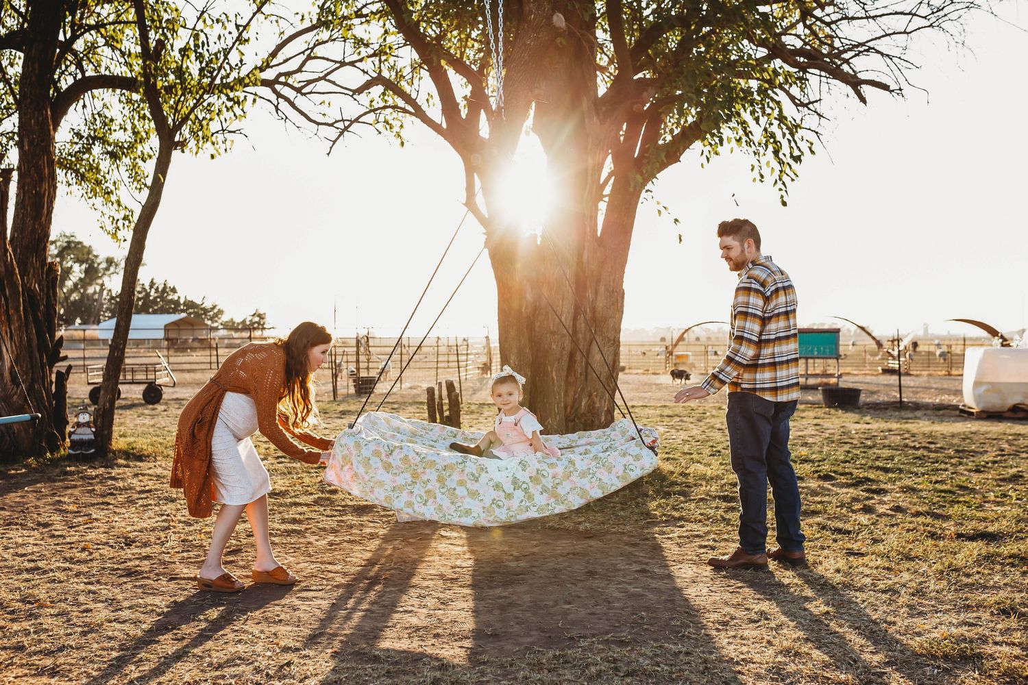 Parents pushing toddler on a hammock swing under a tree.