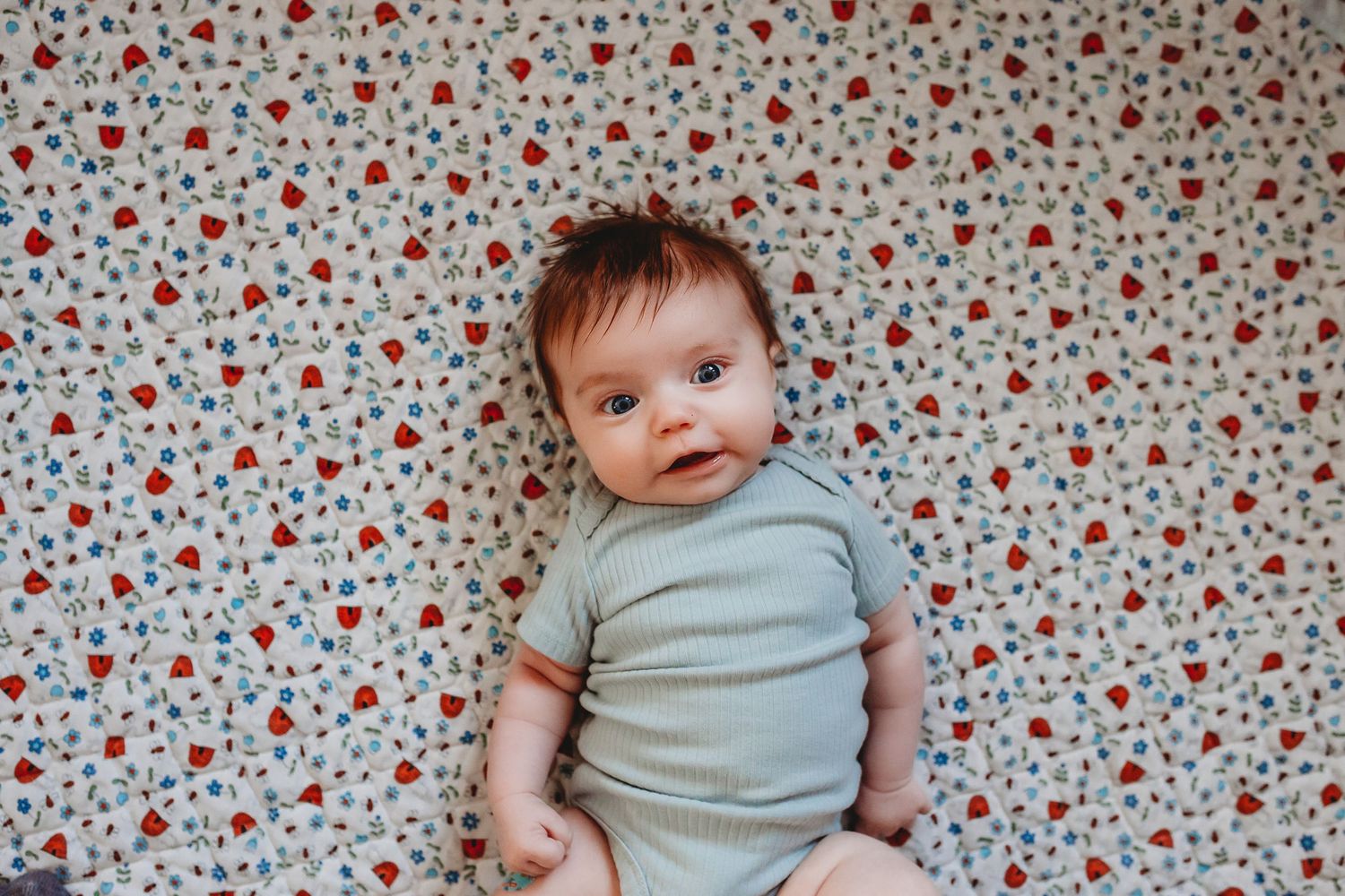 Adorable baby in a teal onesie lying on a strawberry patterned sheet.