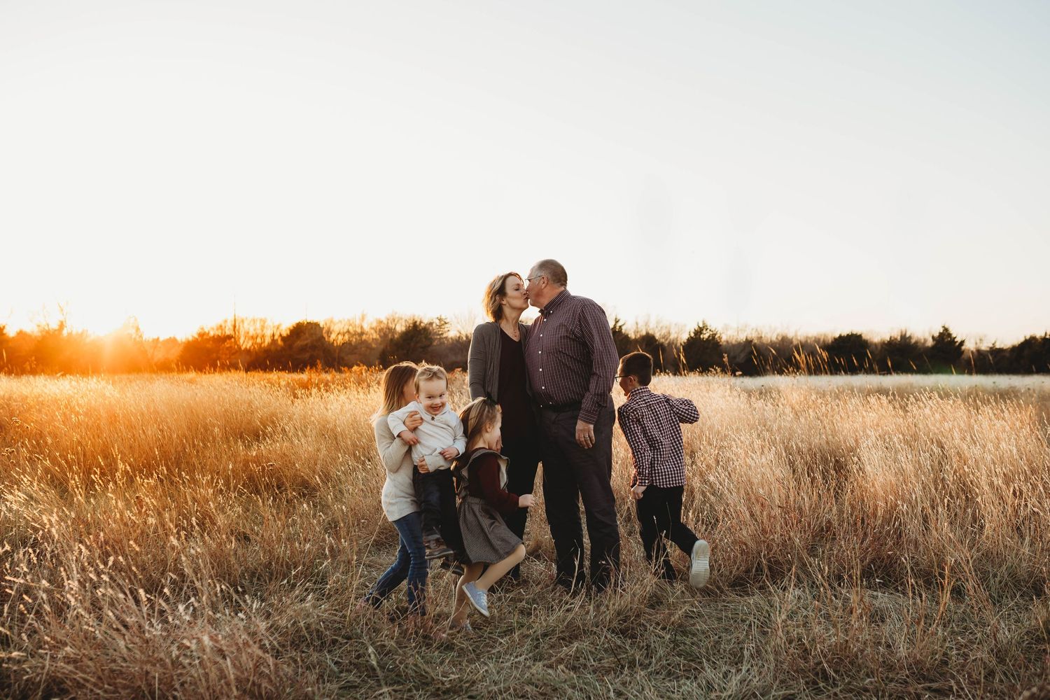 Family of six embracing and kissing in golden tall grass field during sunset.