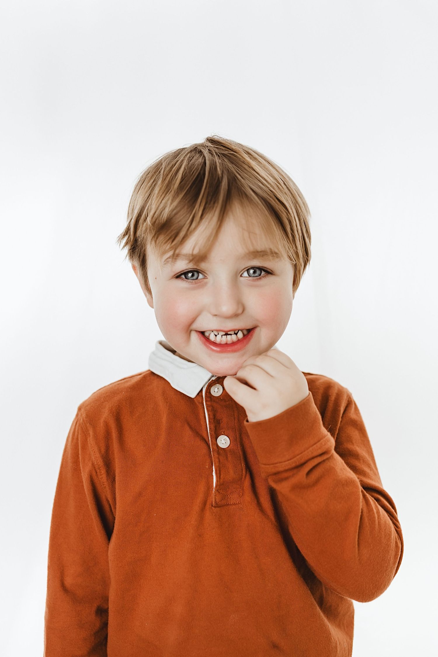 Smiling young boy with blonde hair wearing an orange polo shirt against a white background.