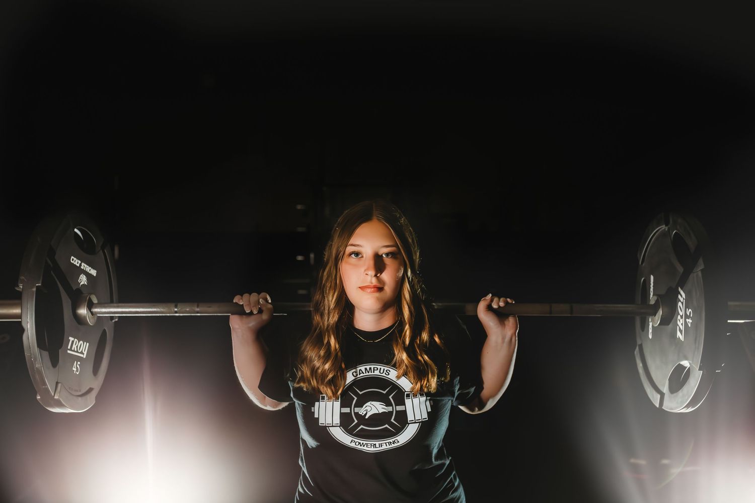 Teen girl confidently lifting a barbell in a dramatic dark studio setting.