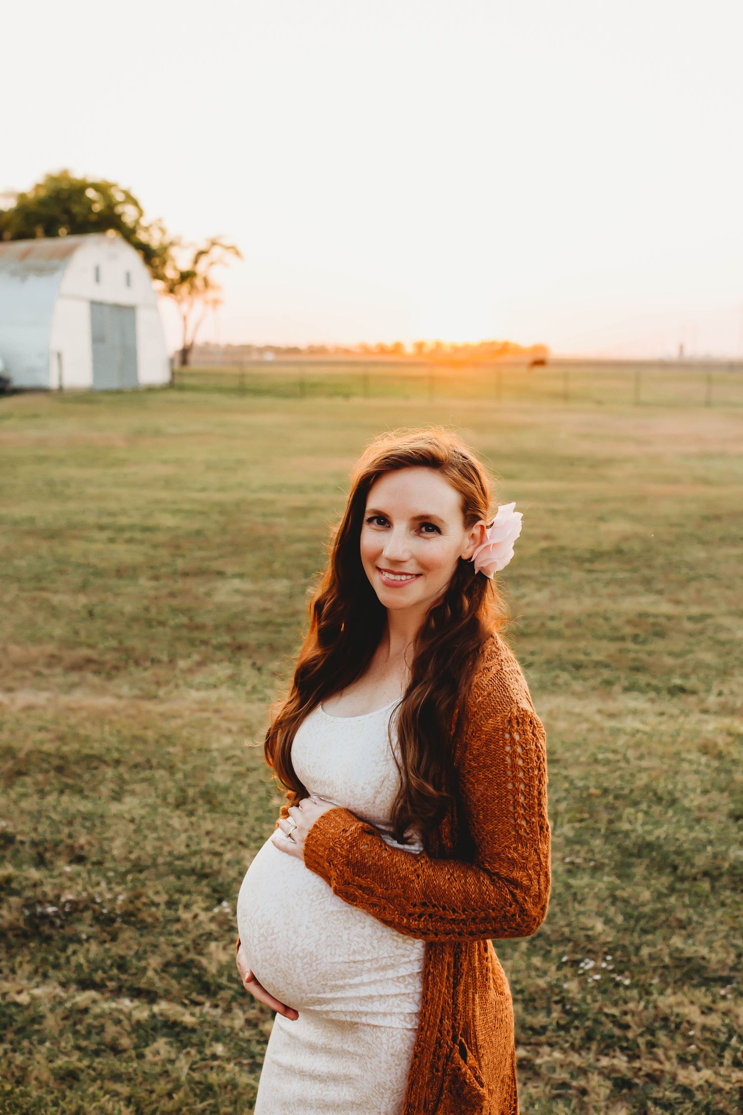 Smiling pregnant woman with flower in hair wearing brown cardigan at sunset near a white barn.