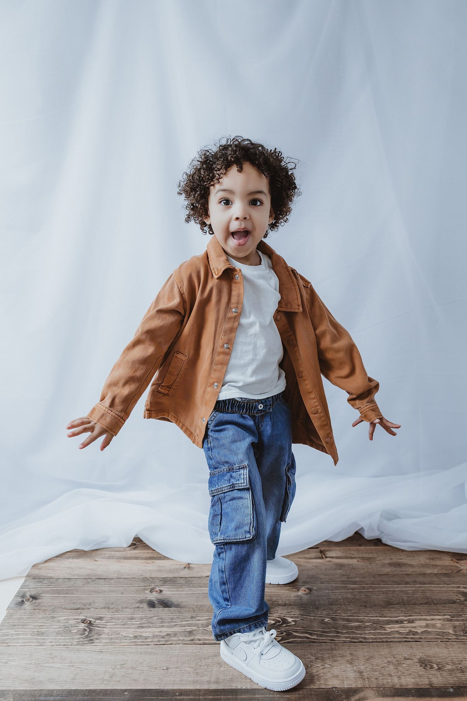 Curly-haired toddler boy in brown shirt and blue cargo jeans posing playfully with tongue out on wooden floor.