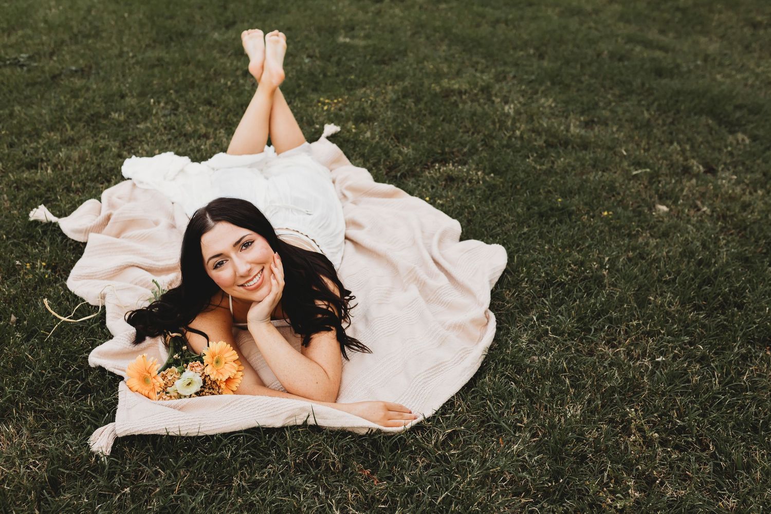 Smiling bride in wedding dress lying on blanket in grass holding bouquet of orange flowers on sunny day.