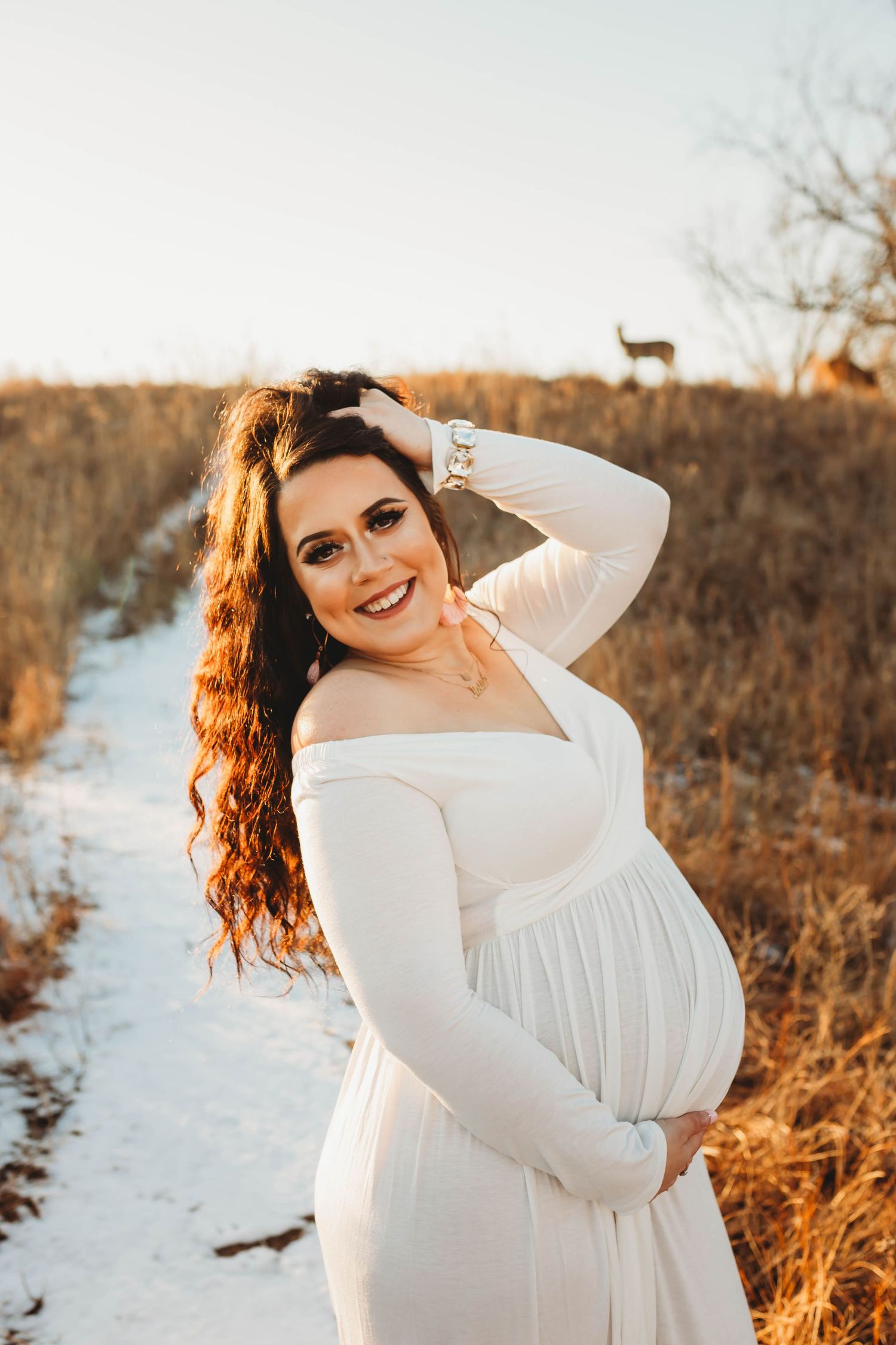 A smiling pregnant woman in a white off-shoulder gown poses on a snowy path at golden hour with deer nearby.