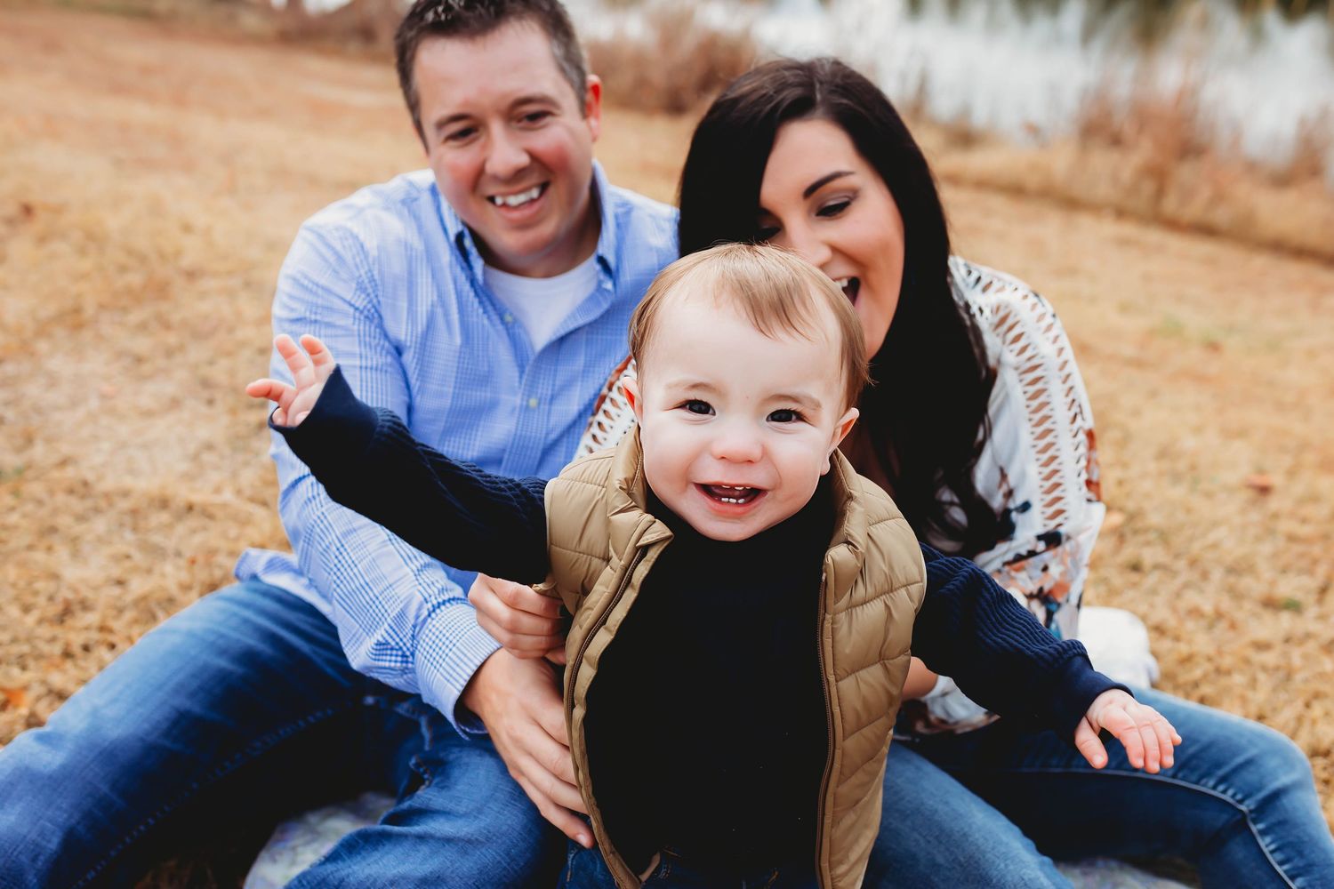 Happy young family with smiling baby sitting together on grass outdoors.