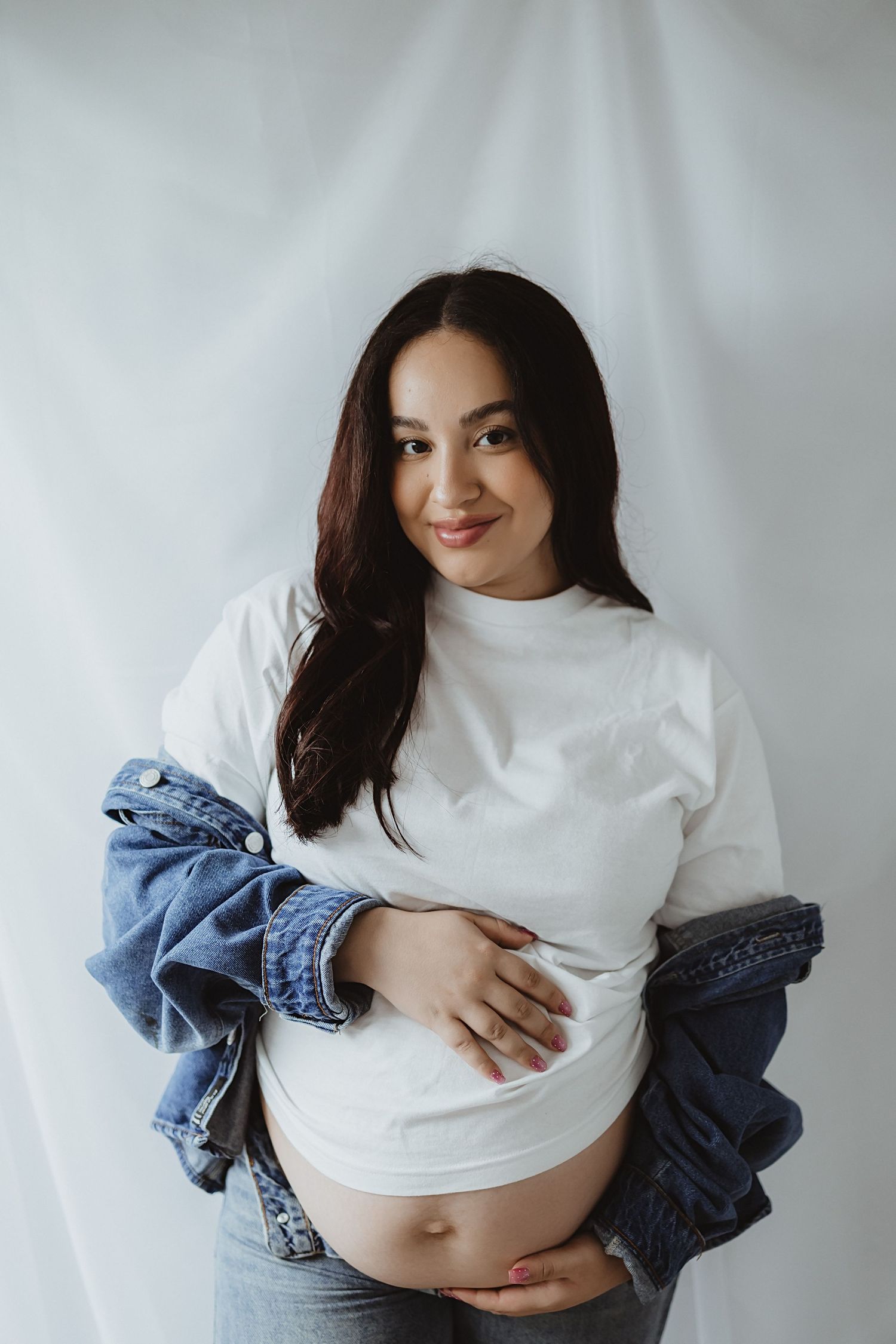 Smiling pregnant woman in white turtleneck and denim jacket gently holds her bare baby bump indoors.