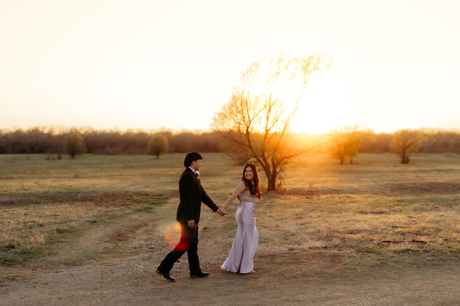 A couple in formal attire holds hands while walking through an open field during golden sunset.