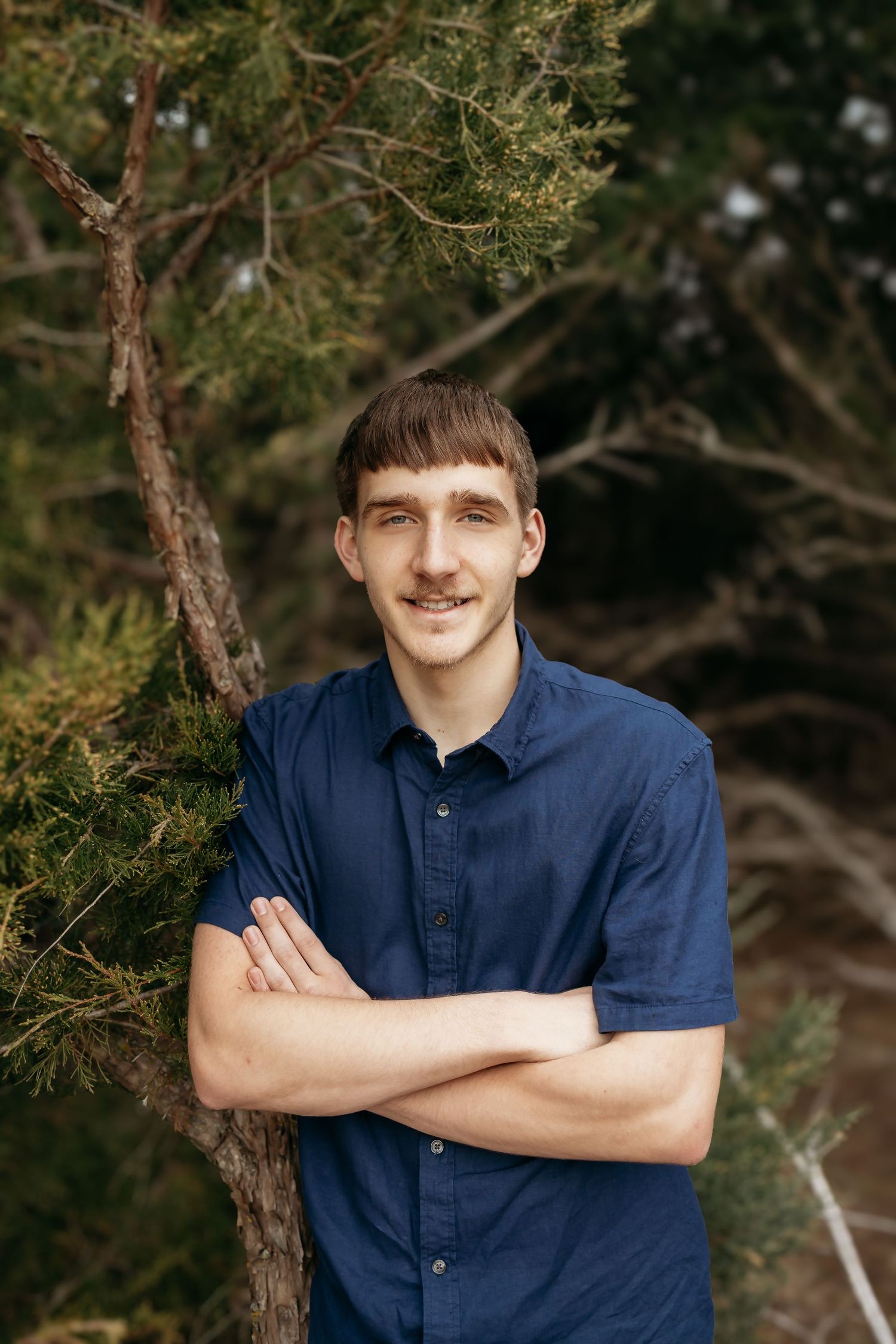 A young man with crossed arms smiles while leaning against a pine tree outdoors.