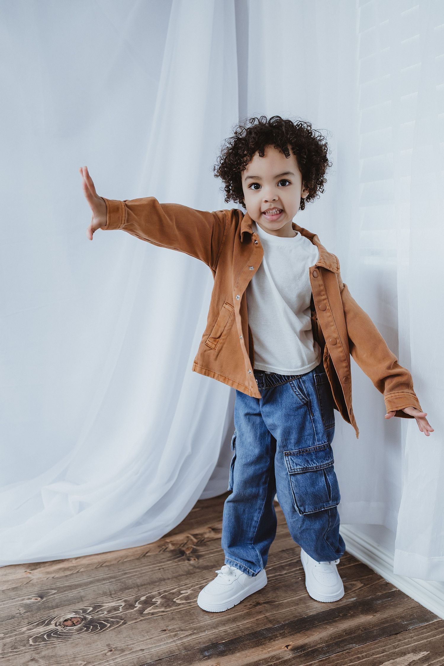 An adorable curly-haired toddler in brown jacket poses playfully with arms outstretched on wooden floor.