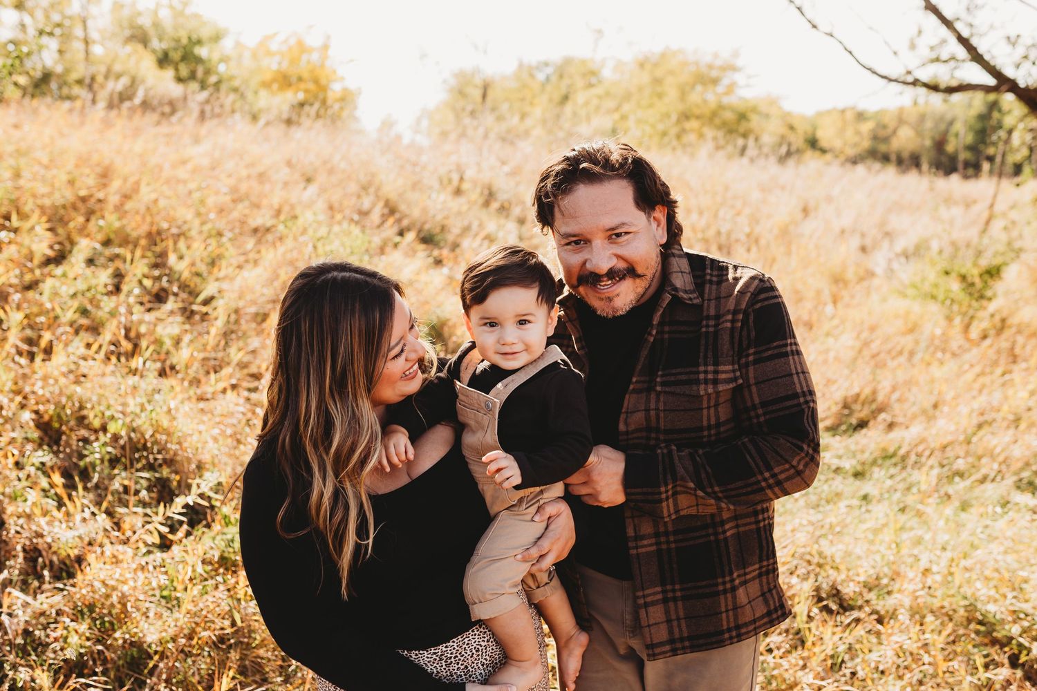 Young couple laughing with their baby boy outdoors in a sunny golden autumn field.