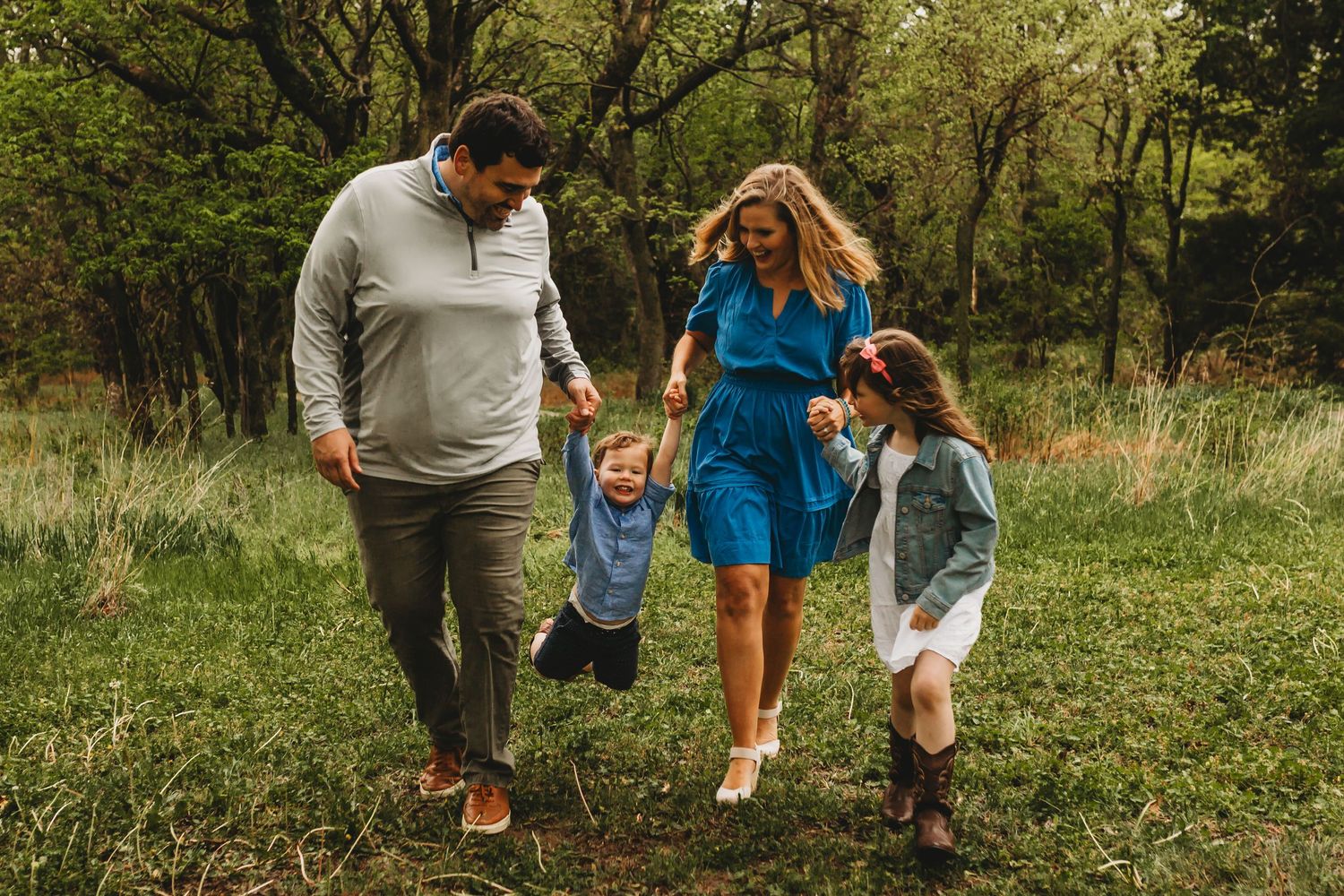 Happy family of four swinging toddler son while walking through lush green meadow together.