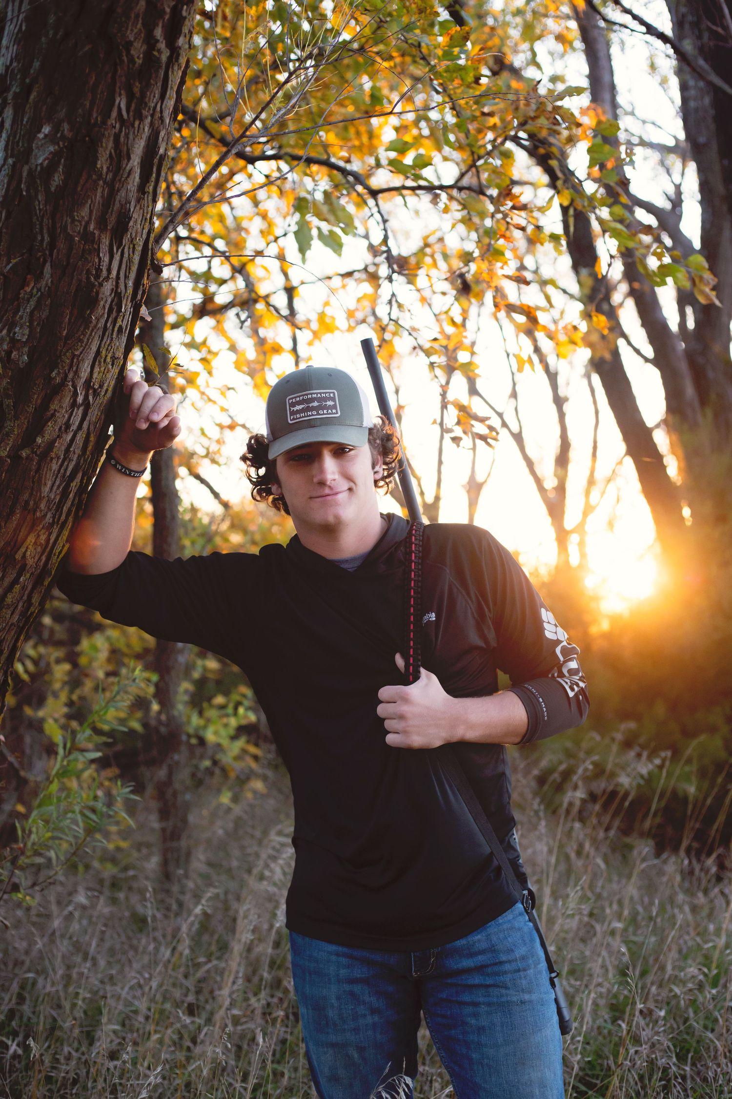 Young man in cap and black shirt leans against tree holding a rifle at golden sunset in autumn forest.