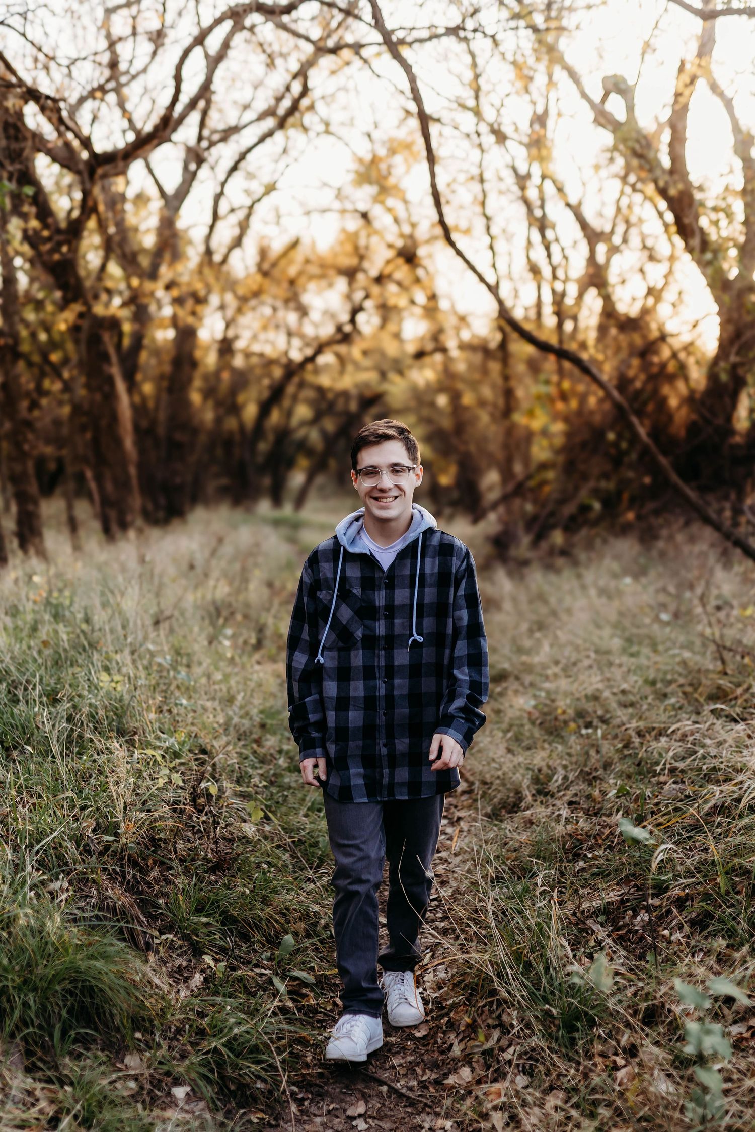 Smiling teenage boy in plaid flannel shirt walking on a forest trail during golden hour autumn sunset.