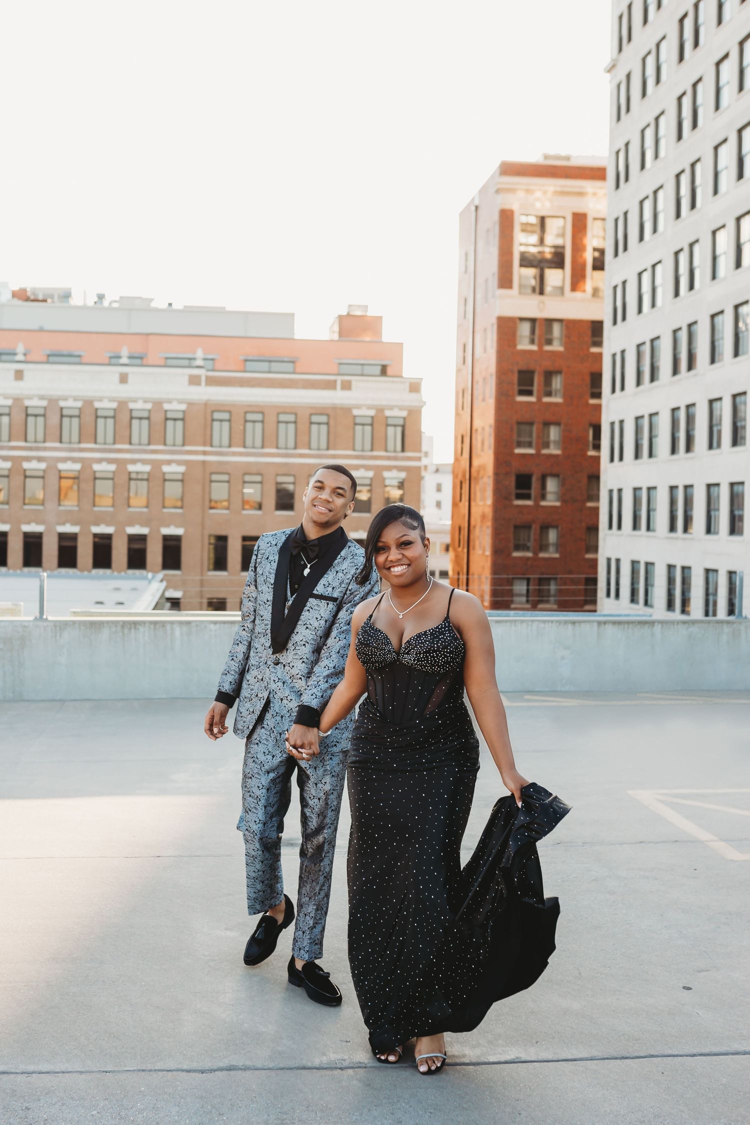 Elegant couple in formal prom attire holding hands on rooftop with city buildings backdrop.