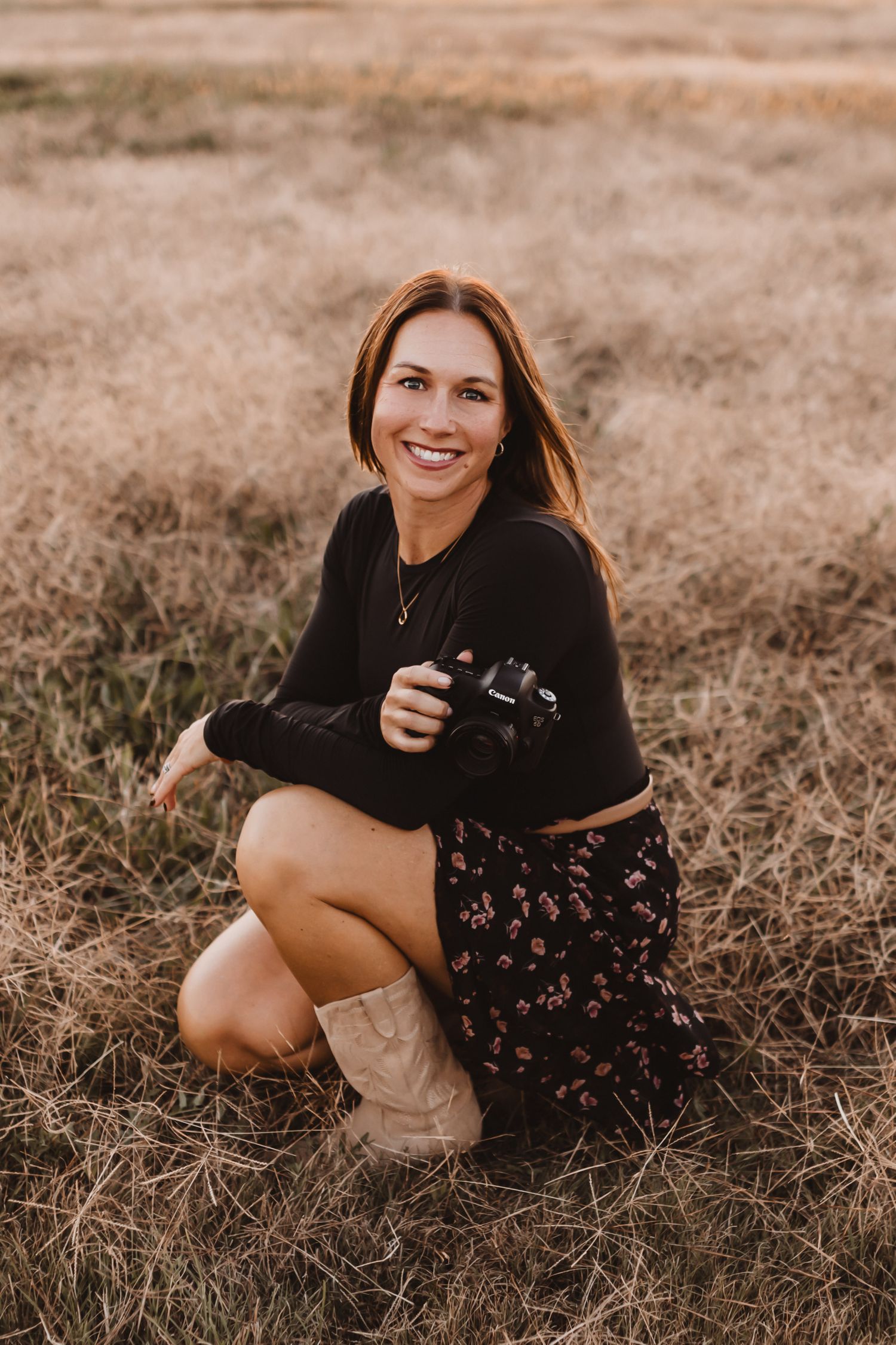 Female photographer smiling and crouching in a golden field holding her camera.