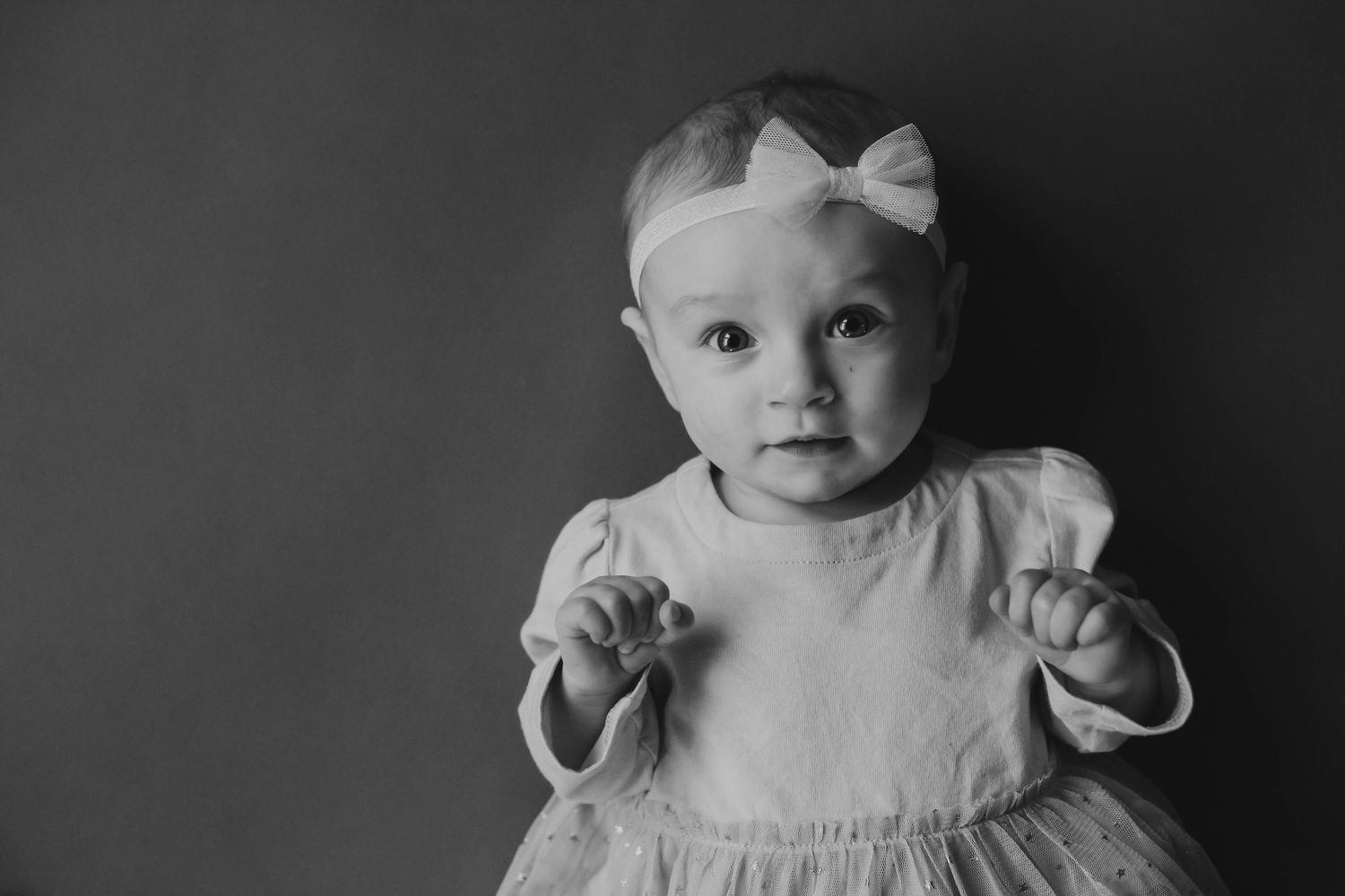 Black and white portrait of an adorable baby girl in a dress with a bow headband looking at the camera.