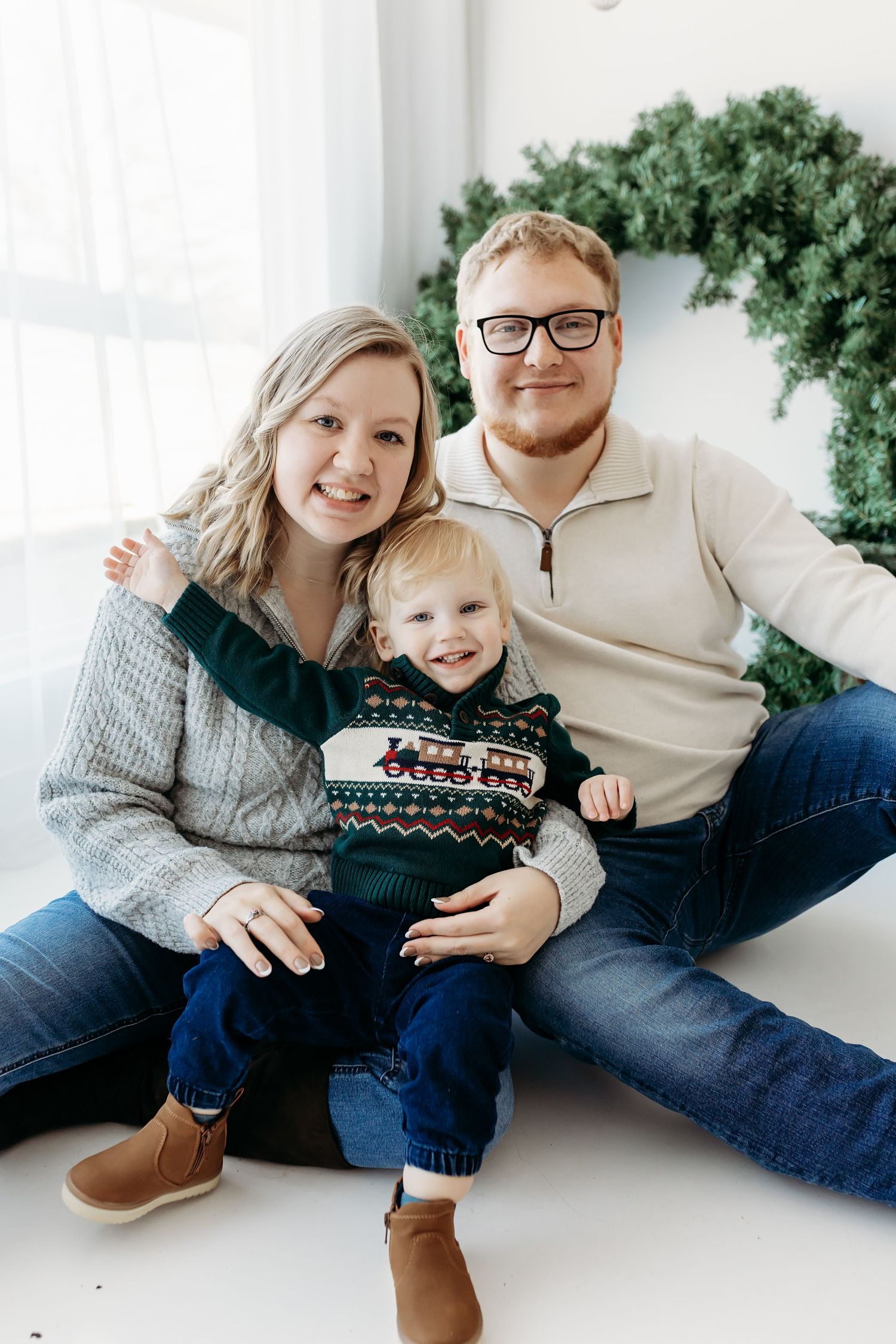 A smiling young family of three poses together in front of a Christmas wreath in a bright studio.