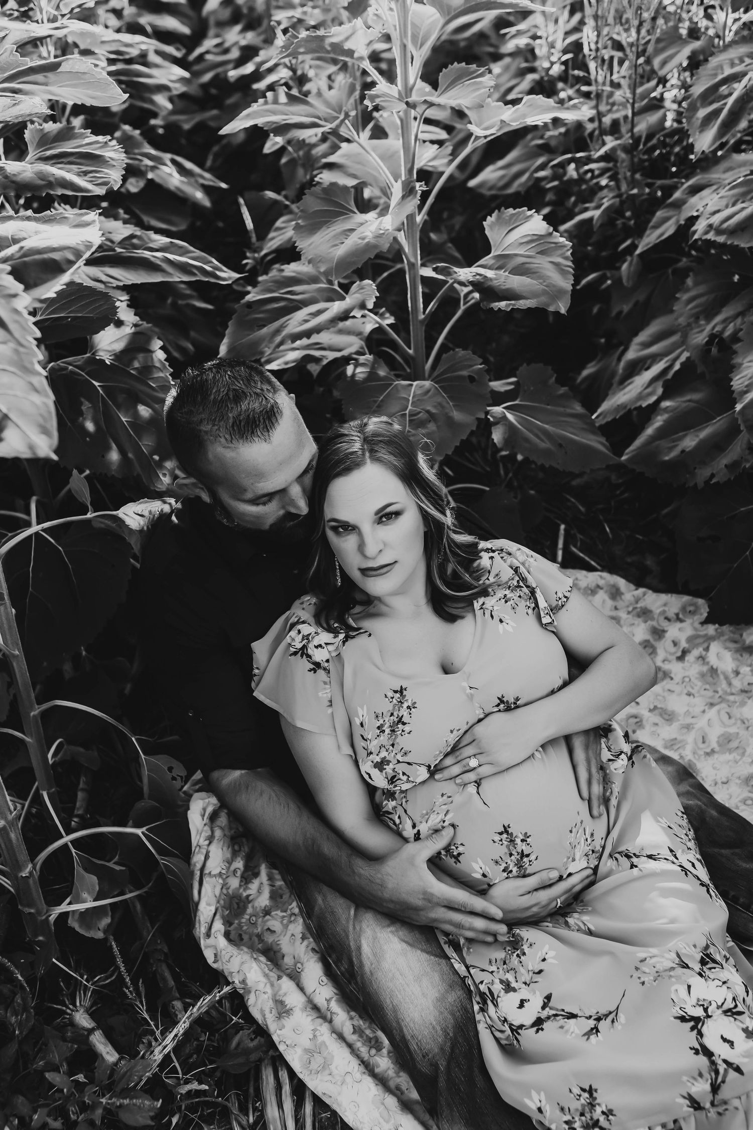 Black and white photo of a couple sitting among lush tropical plants, tenderly cradling a pregnancy bump.