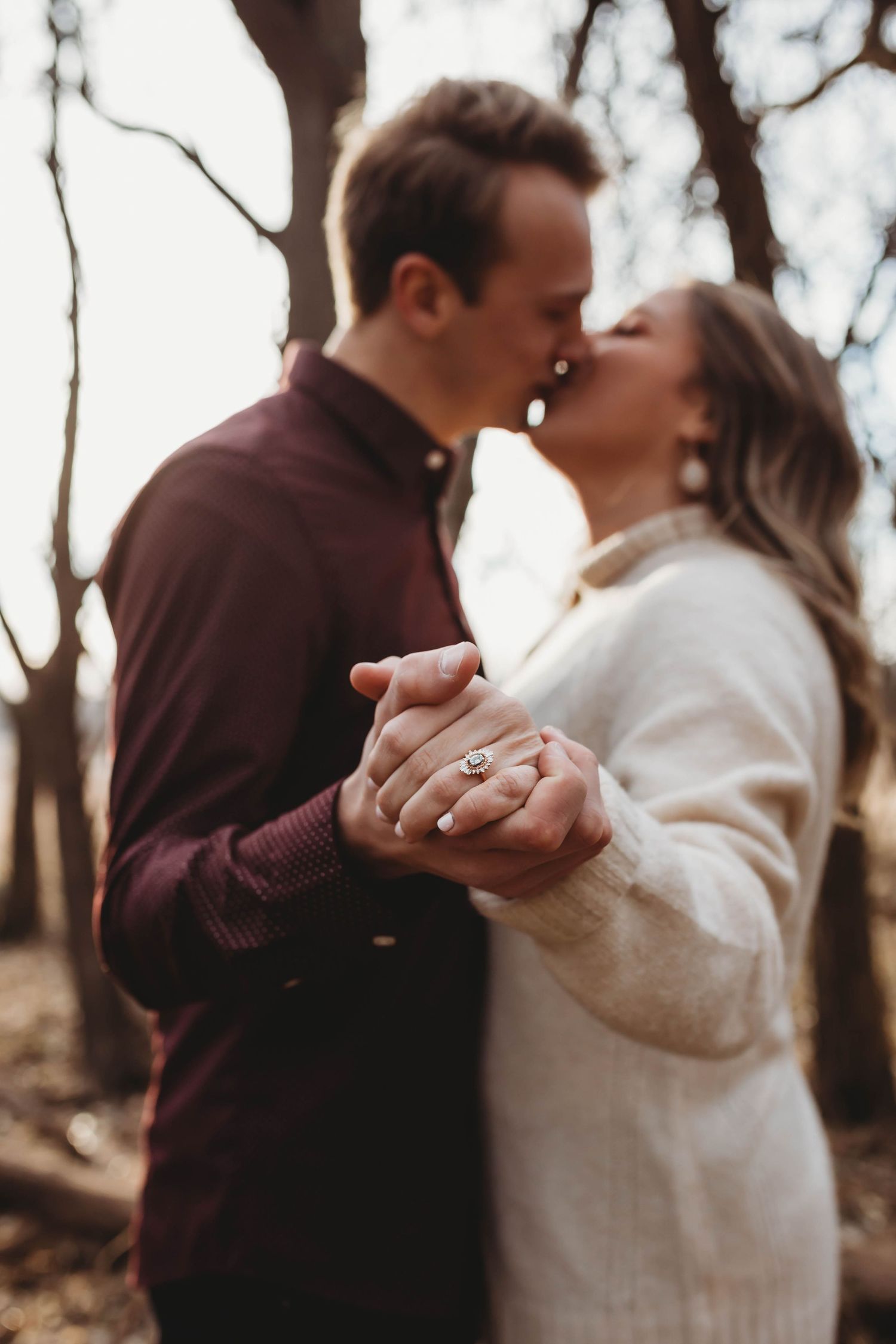 Engaged couple kissing while holding hands showing diamond engagement ring in a romantic forest setting.