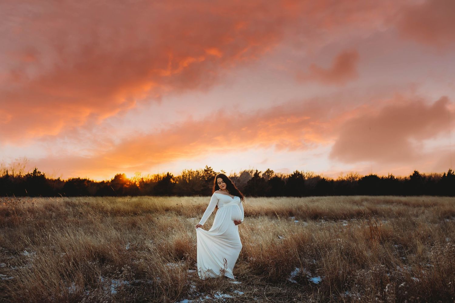 Woman in white dress walking through golden field at dramatic sunset with vibrant orange and pink sky.
