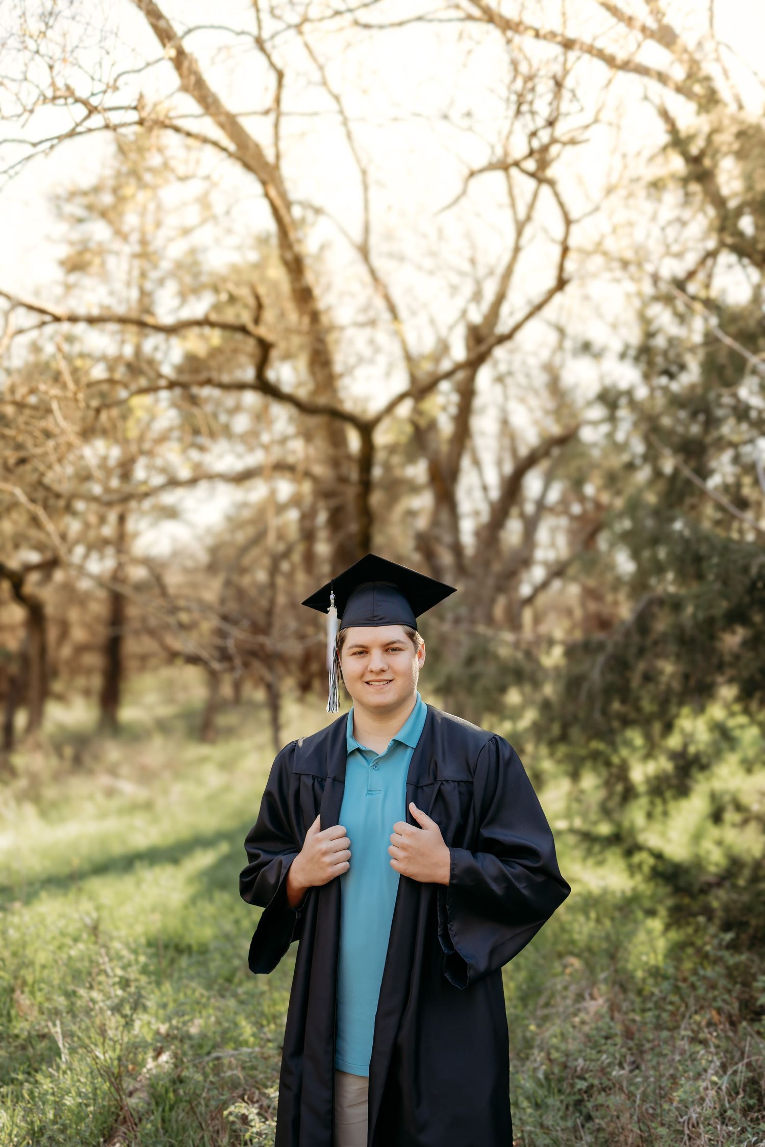 A smiling young man in graduation cap and gown stands outdoors among autumn trees at sunset.