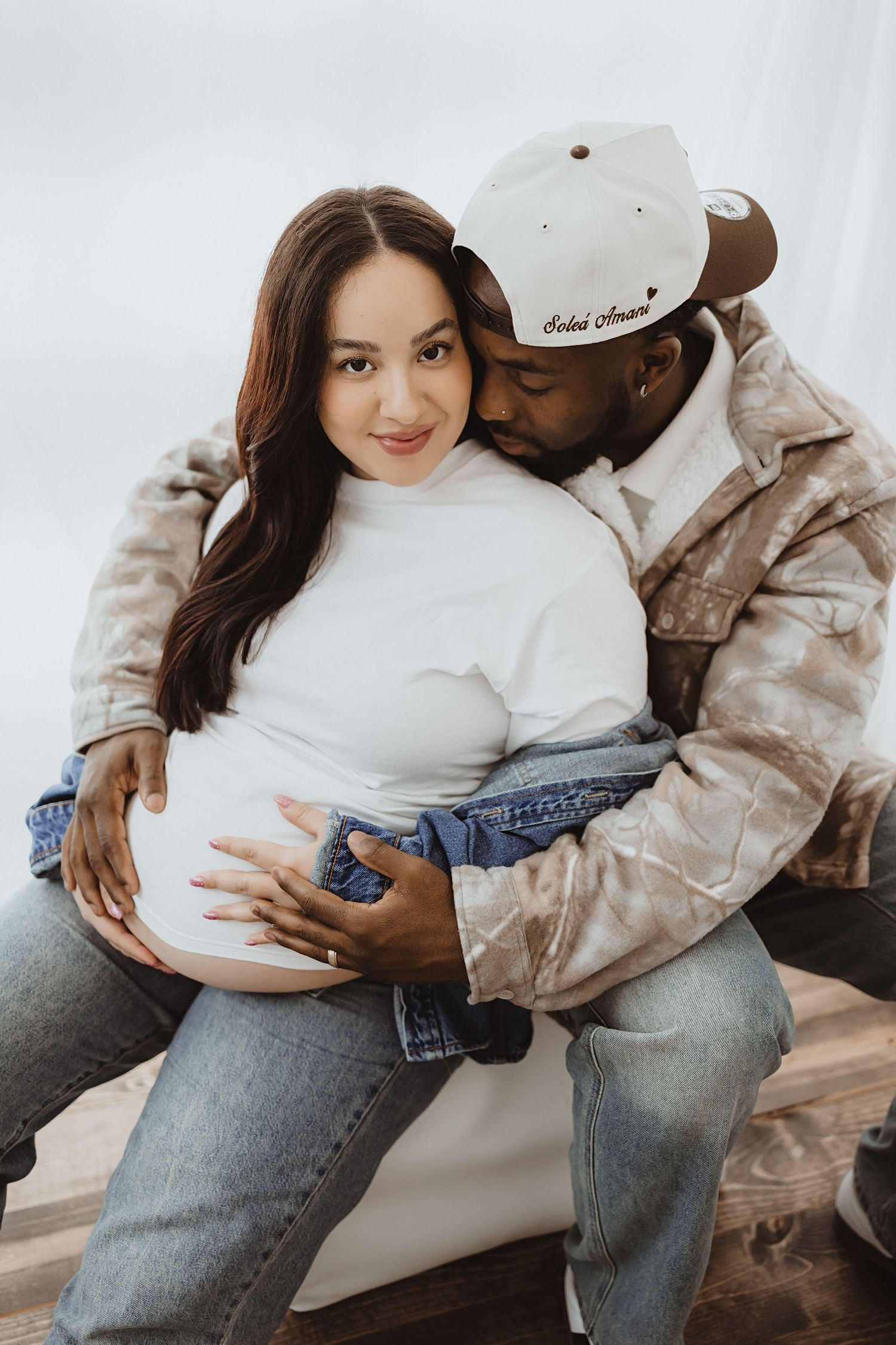 Interracial couple shares tender maternity moment, man in cap embraces pregnant woman from behind holding her belly.
