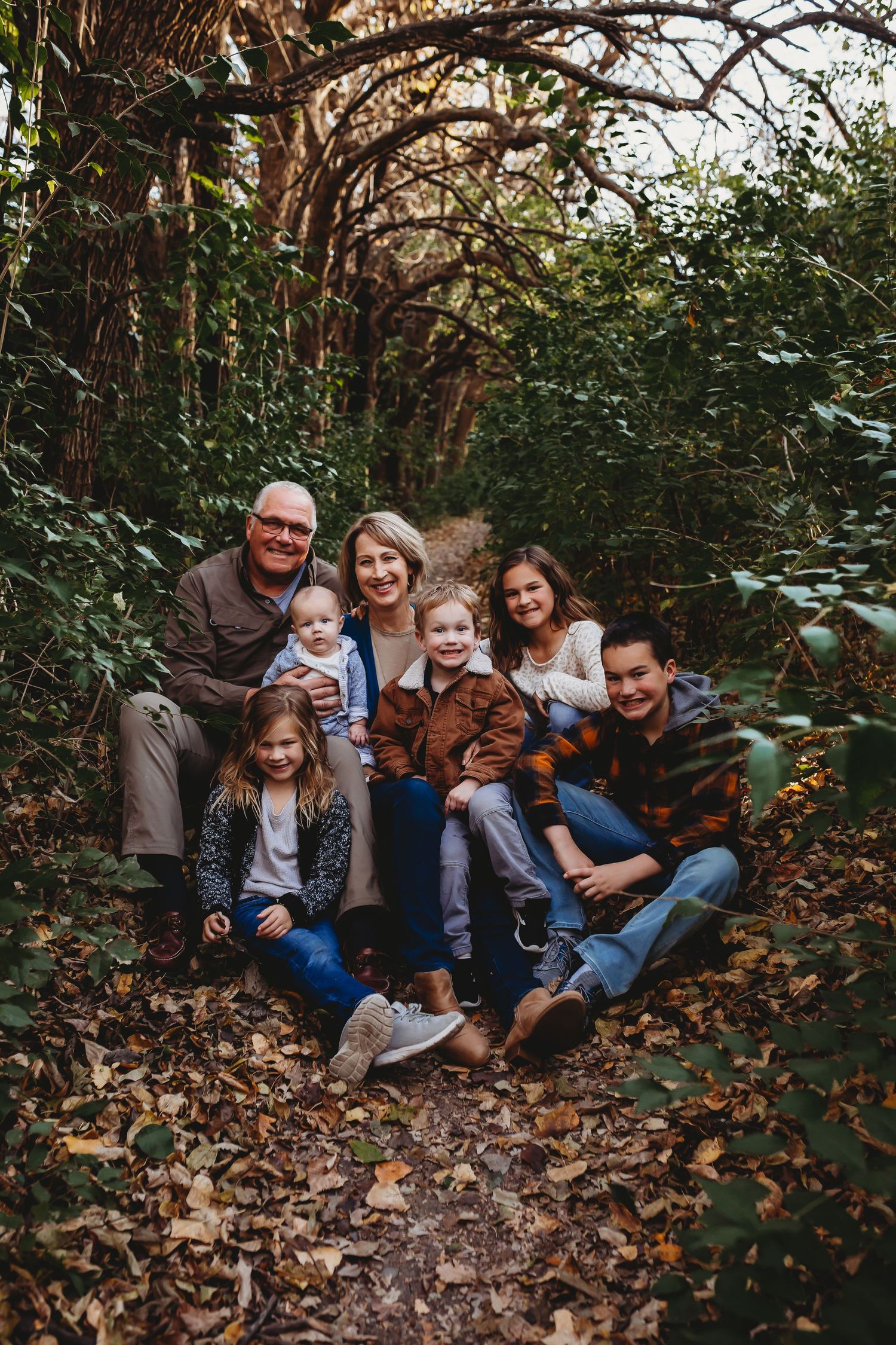 Extended family portrait sitting on fallen leaves in a lush green forest tunnel.