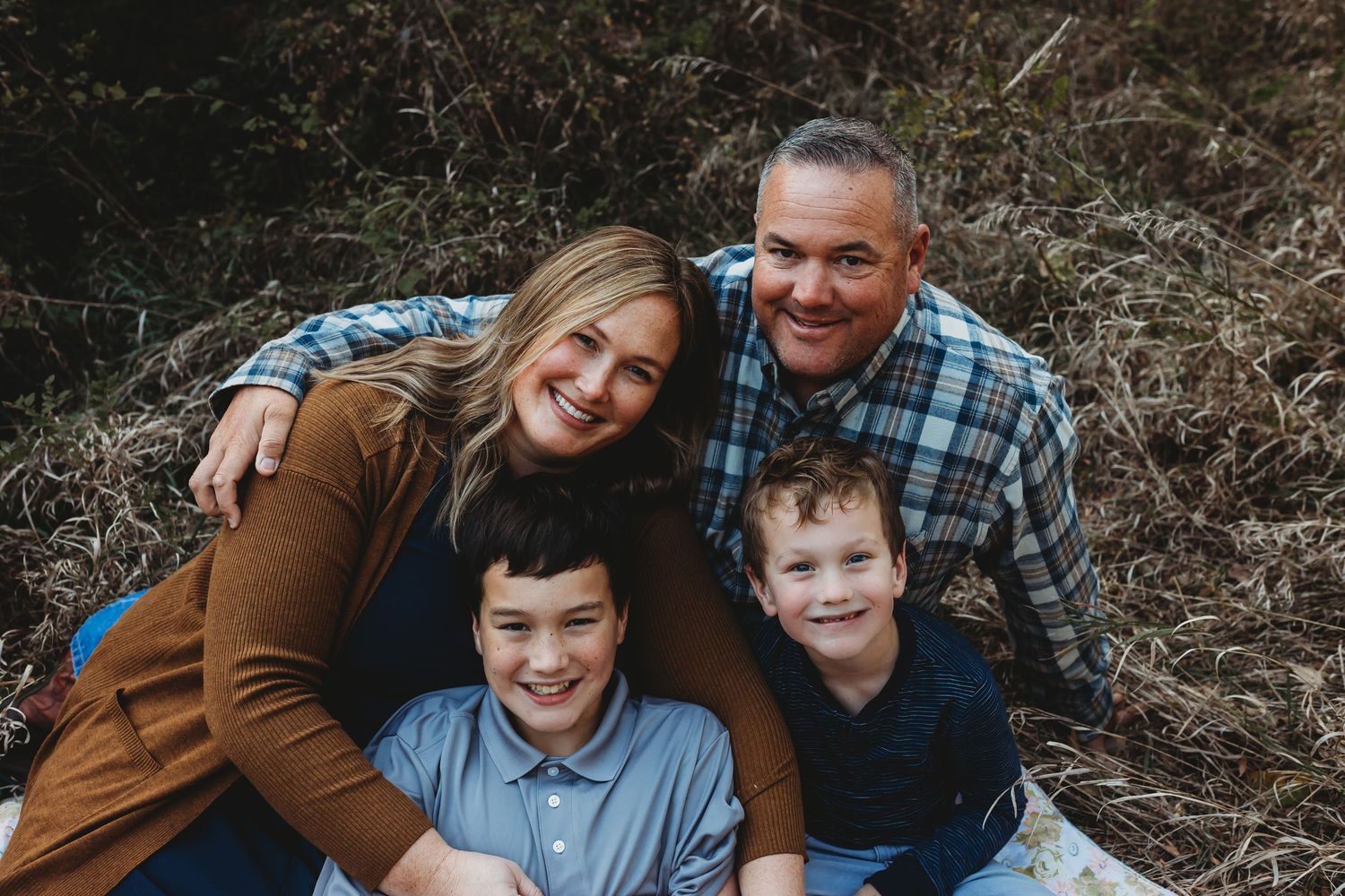 Family of four smiling together on a blanket in nature.