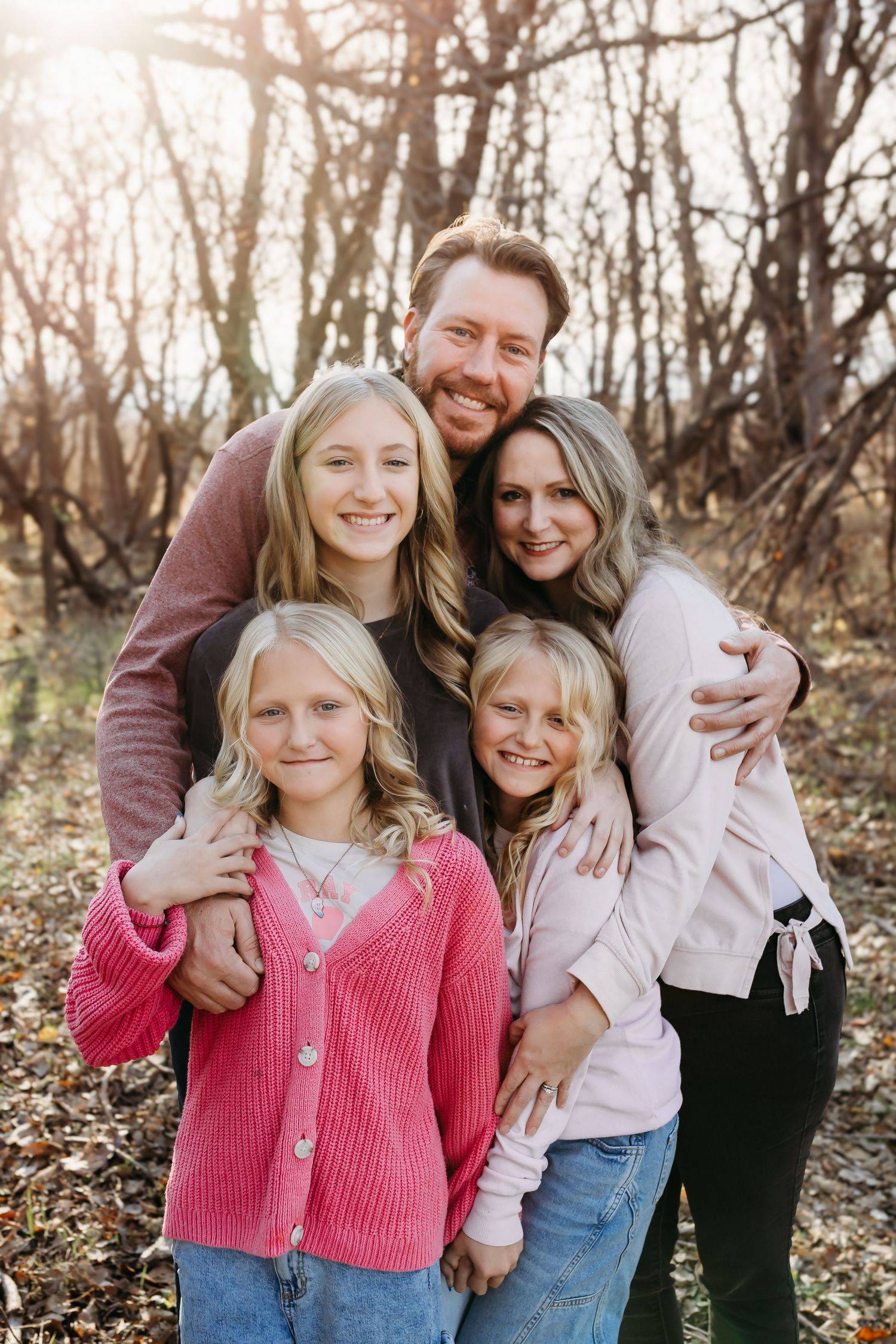 Family of five hugging closely outdoors in winter woods, wearing coordinating pink and maroon outfits.