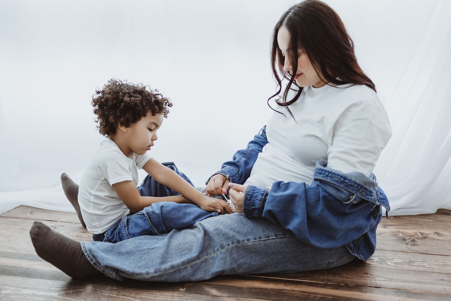 Pregnant woman in white top and jeans sits on wooden floor with curly-haired toddler during maternity photo session.