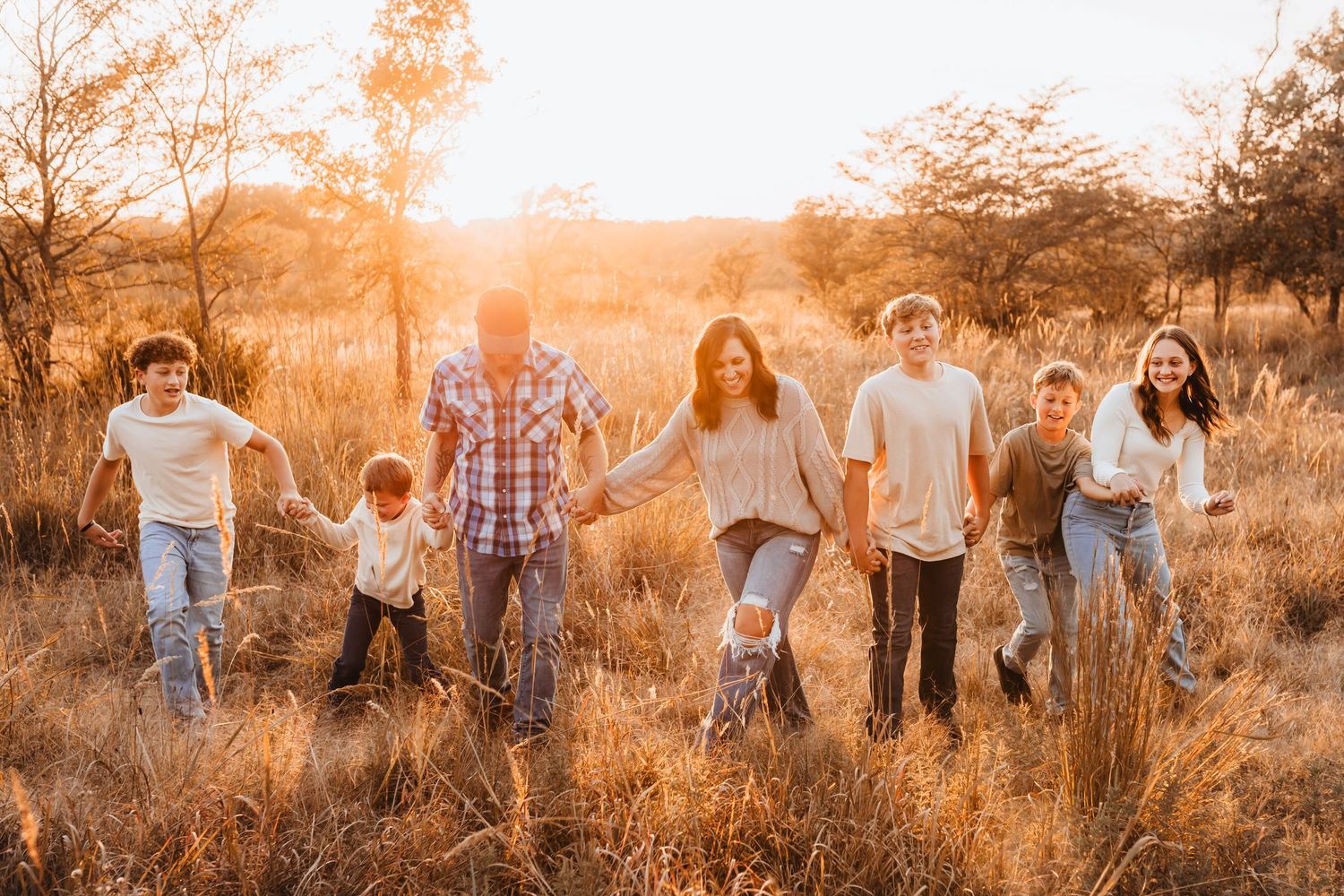 Family of seven holding hands walking through golden field at sunset in warm autumn light.
