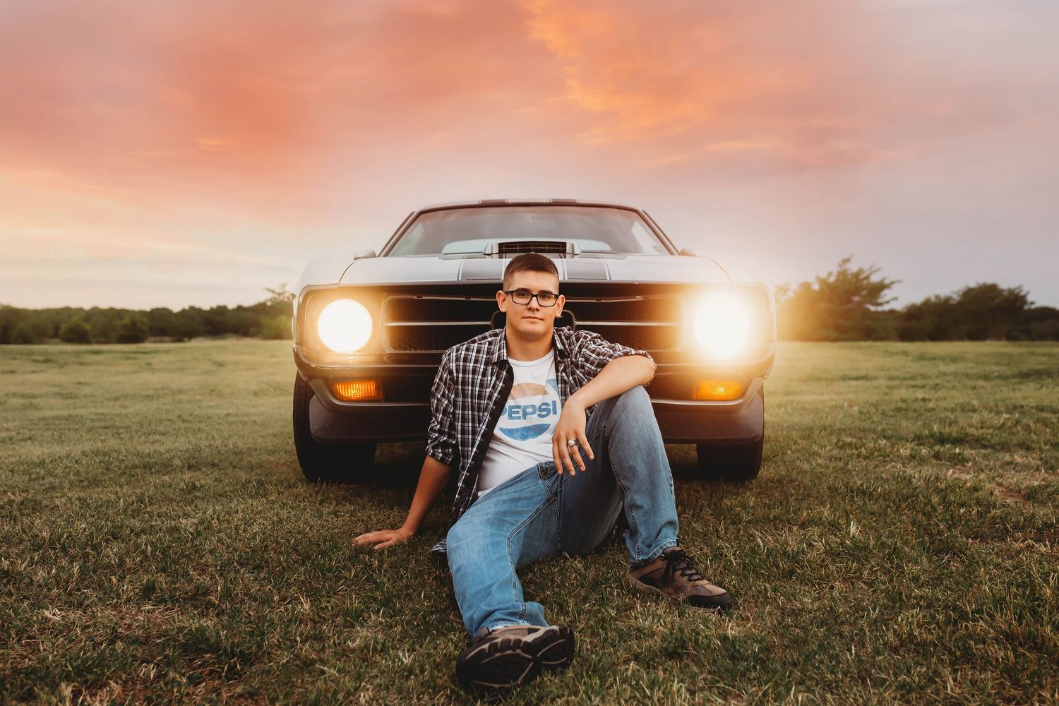 Teen boy sitting in front of classic black muscle car at colorful sunset outdoors.