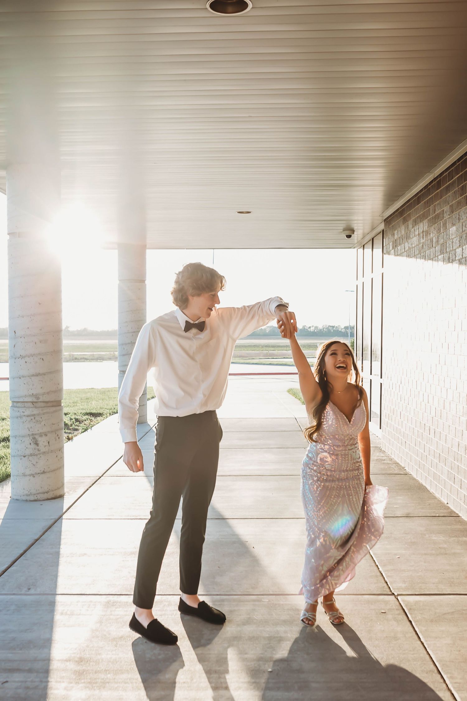 Couple in formal attire dancing joyfully in a sunlit corridor, man in white shirt and bow tie twirling woman in sequin dress.
