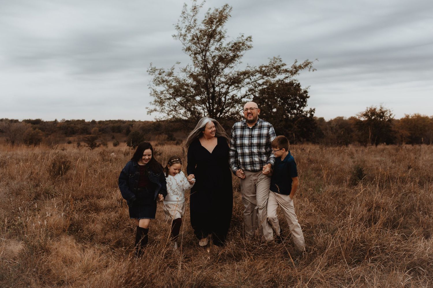 Family of five posing in an open field with bare trees and overcast skies during an outdoor fall photo session.